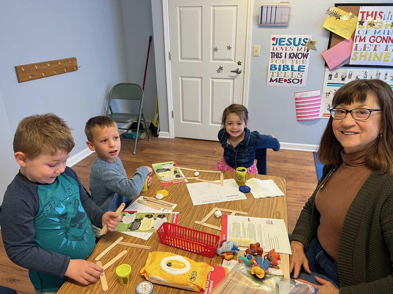 A smiling woman with glasses sitting at a table with three young children, engaging in arts and crafts; the table has toys and craft supplies, and the room has decorated walls with colorful posters and children's artwork.
