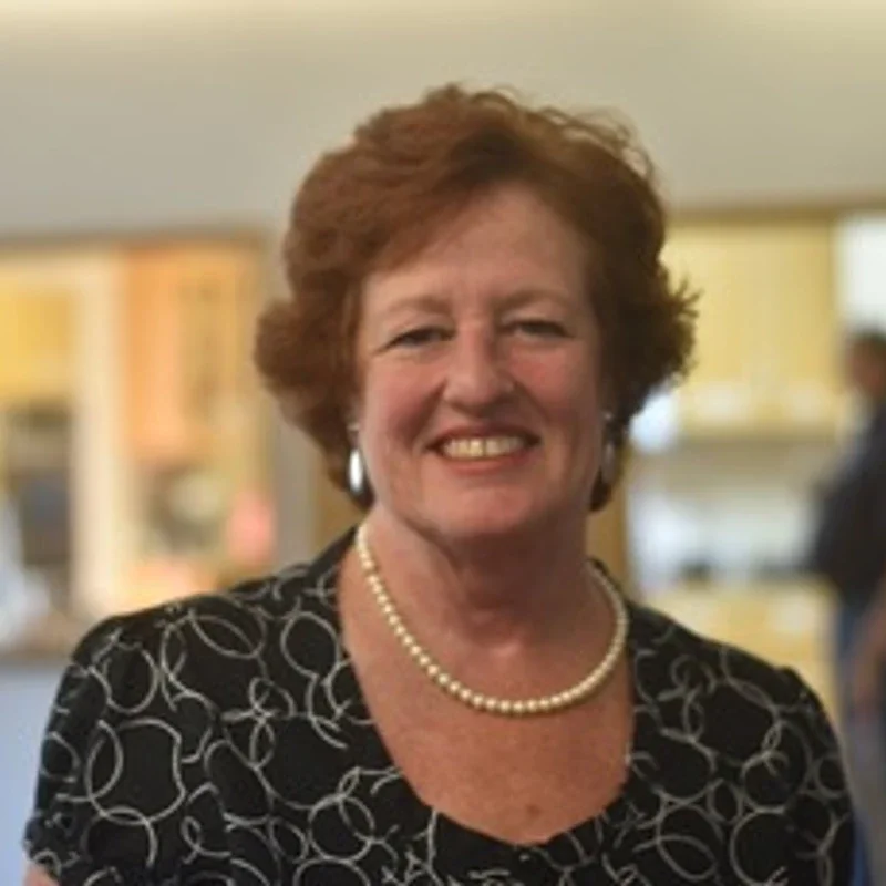 Smiling woman with short red hair, wearing pearl necklace and patterned black blouse, in an indoor setting.