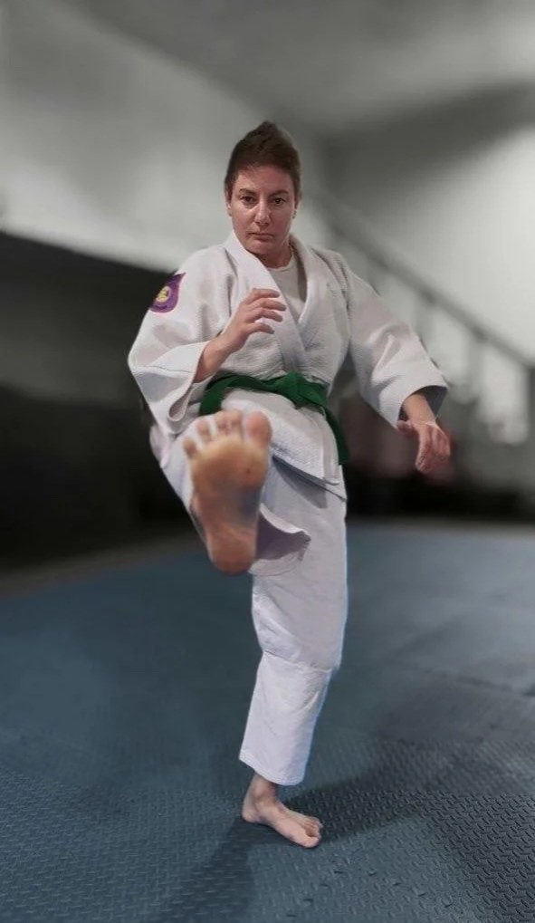 Bukan instructor Ioana Florescu in a white karate gi with a green belt is performing a kick in the dojo.