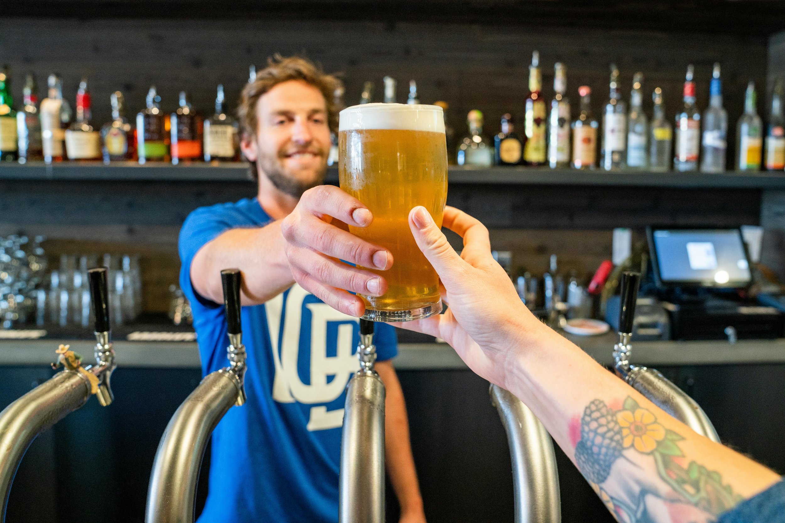 A person with tattoos on their arm is holding a glass of beer, clinking it with a bartender behind the bar. The bartender is smiling and wearing a blue shirt.