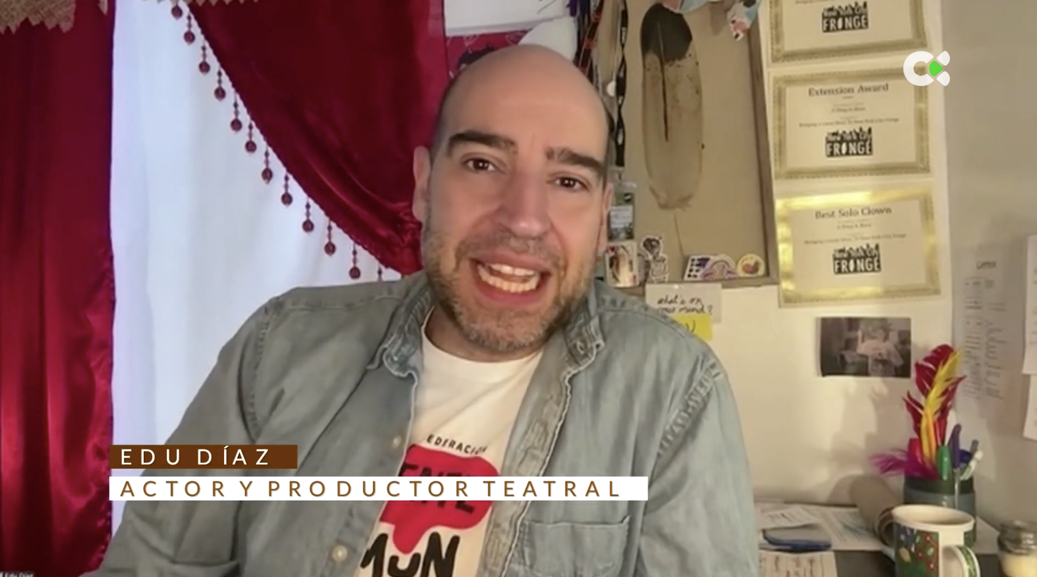Edu Díaz. A man with a beard and bald head smiling at the camera, sitting at a desk with colorful pens, papers, and awards on the wall behind him, in a room with red curtains.