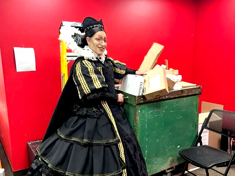 Edu Díaz in PETRUS. A person dressed in a black and gold costume with a ruffled collar, standing next to a green recycling bin filled with cardboard boxes, against a red wall.