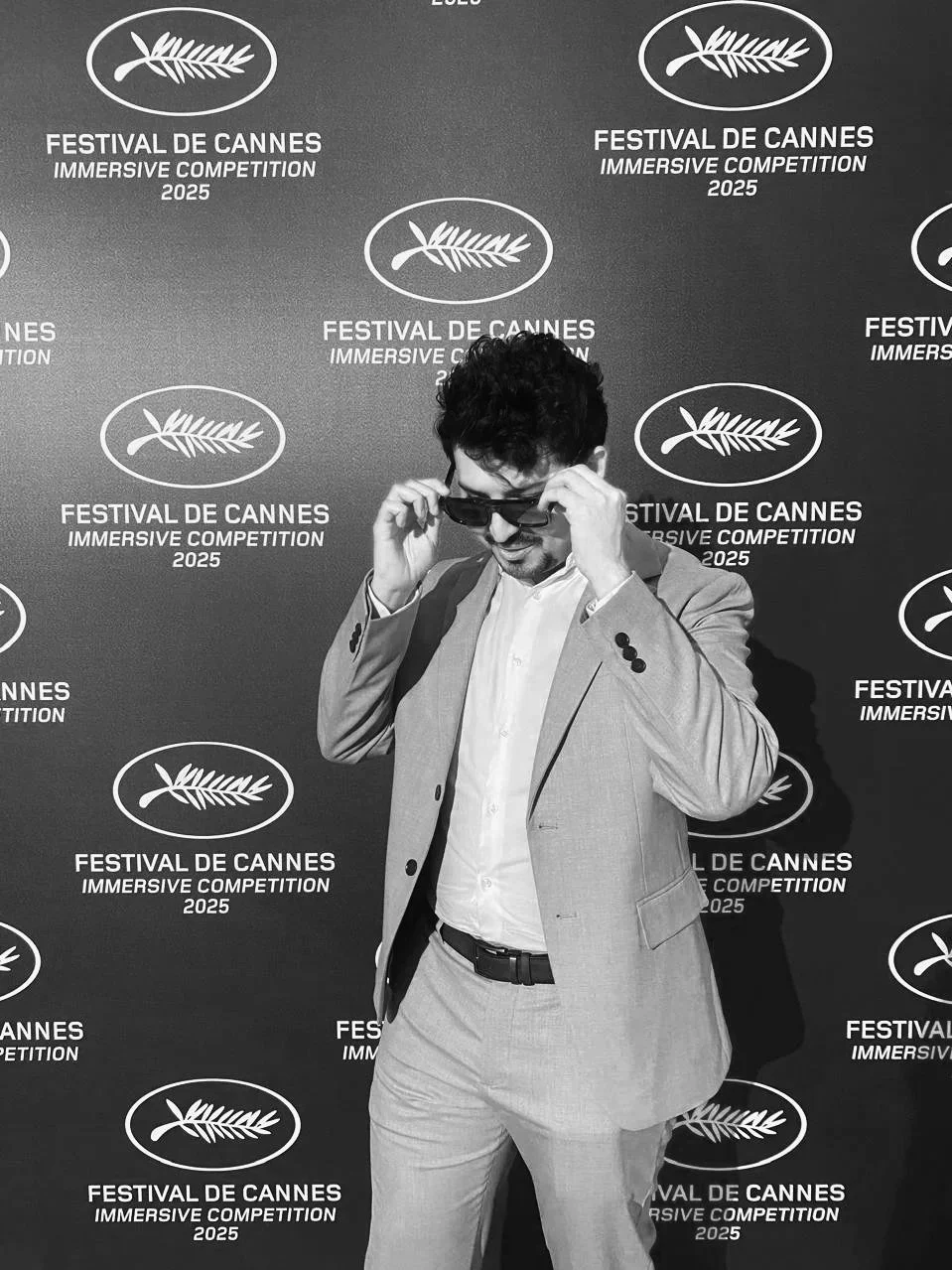A man in a light suit and sunglasses adjusts his glasses while smiling at the Cannes Film Festival 2025, standing in front of a backdrop with the Cannes logo and event details.
