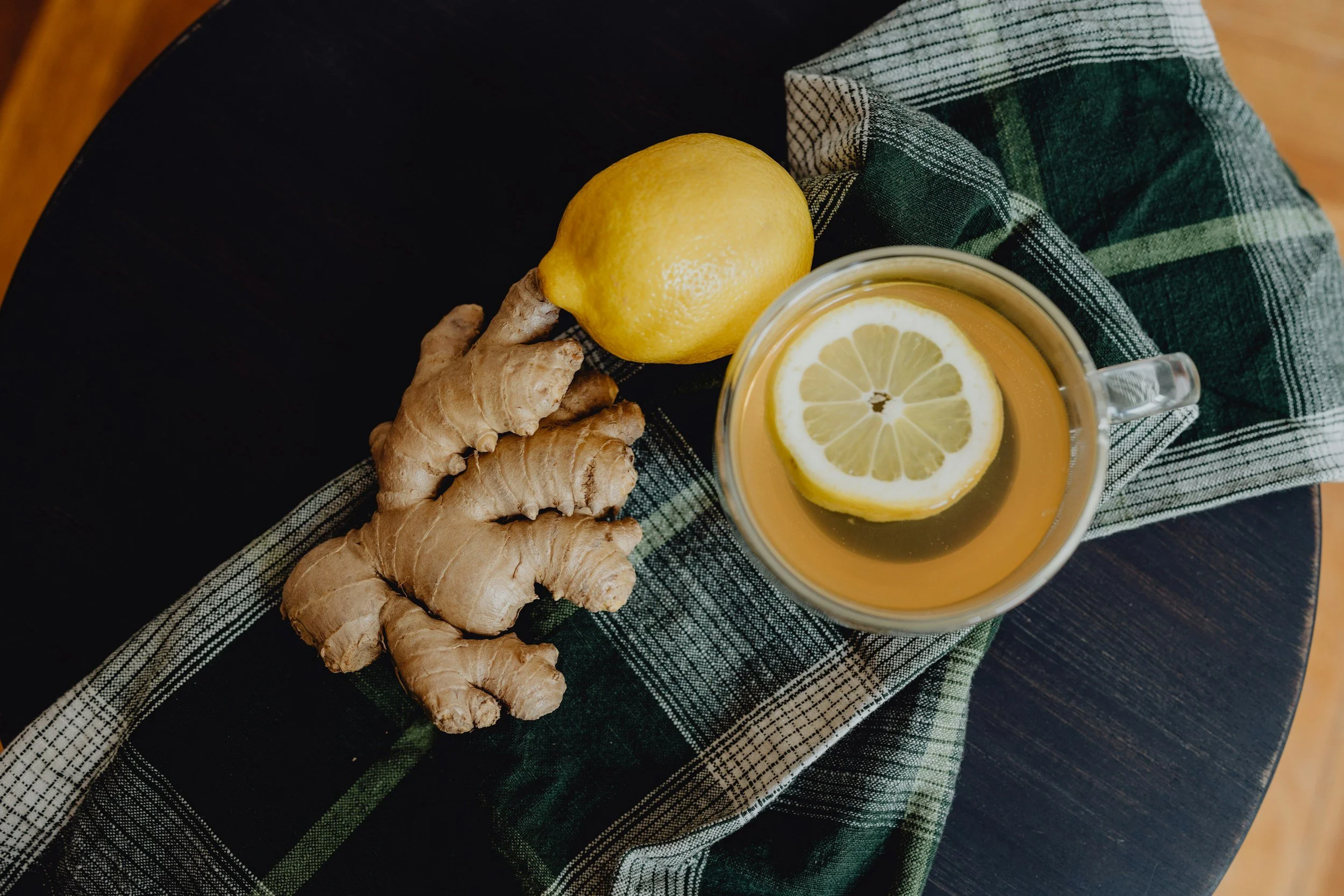 A lemon, ginger root, and a mug of tea with a lemon slice on top are placed on a green checkered cloth on a dark wooden table.