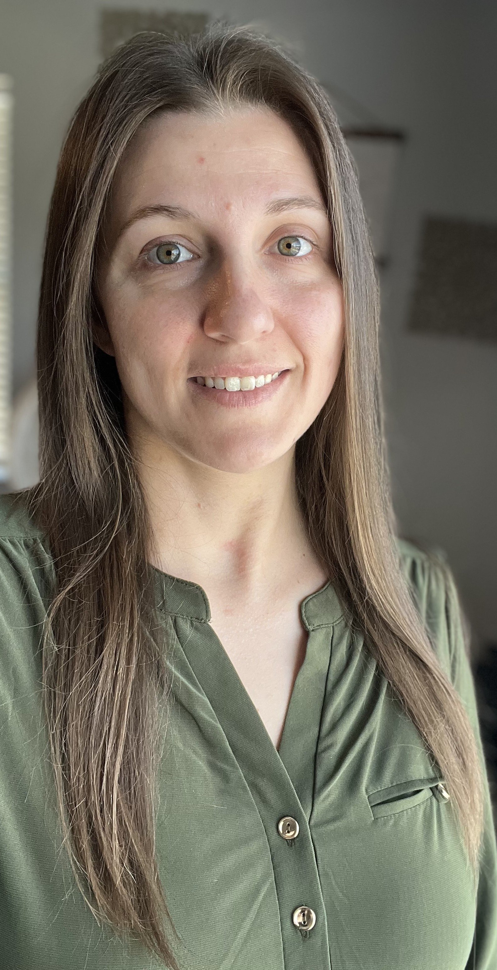 Close-up of a woman with long brown hair, green eyes, wearing a green button-up shirt, smiling indoors.