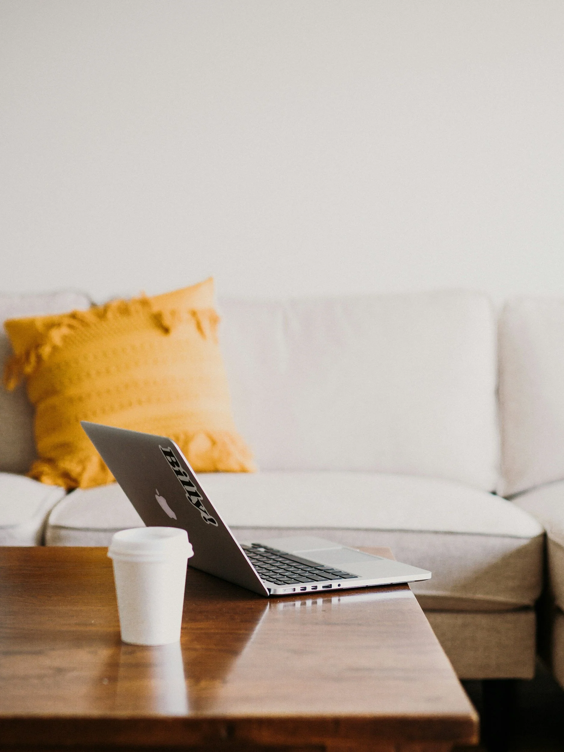 Open laptop on wooden table with coffee cup in foreground, behind it a yellow pillow on a white sofa and a white wall.