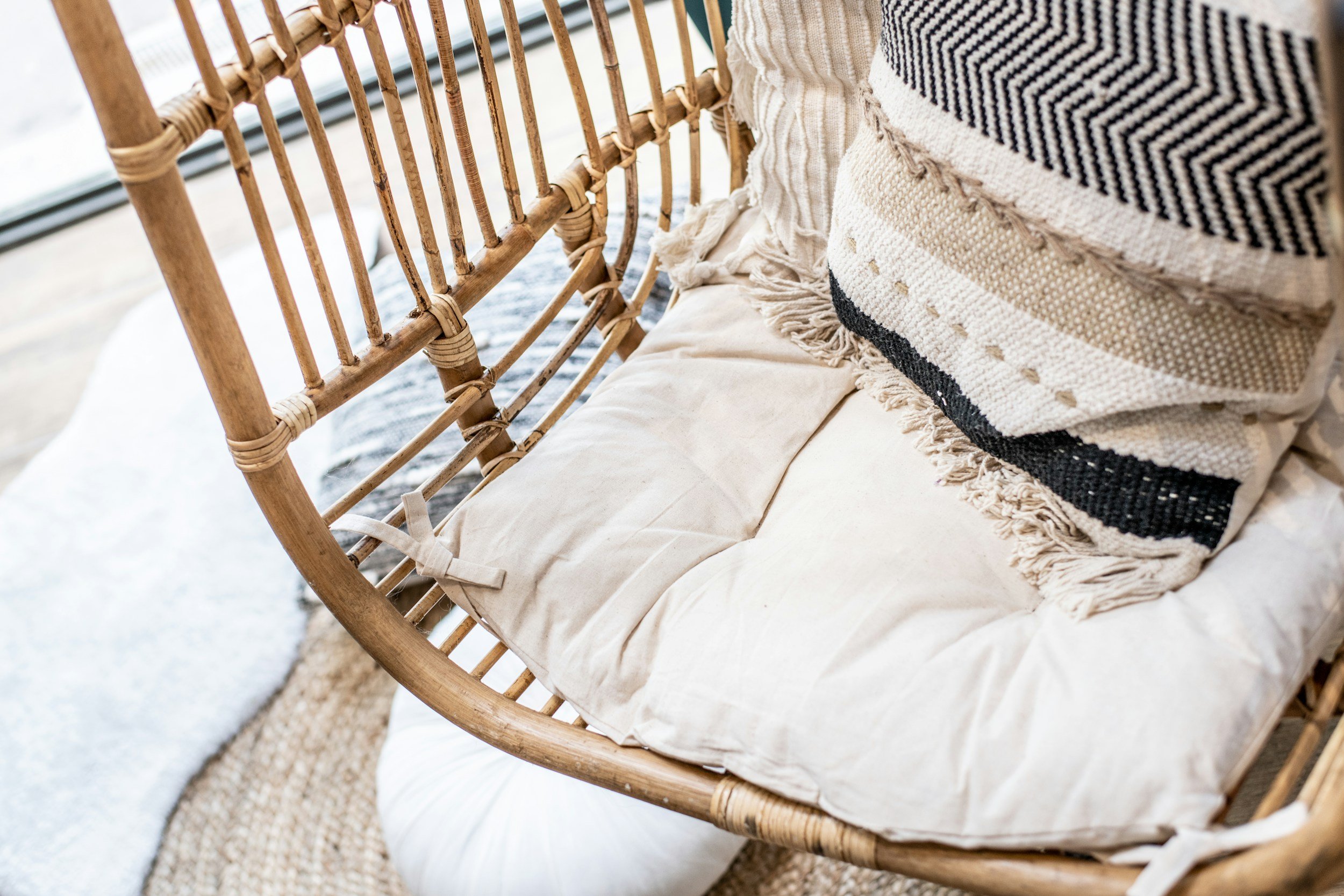 Close-up of a rattan outdoor sofa with neutral-colored cushions, a woven throw blanket, and decorative pillows, situated near a window with a view outside.