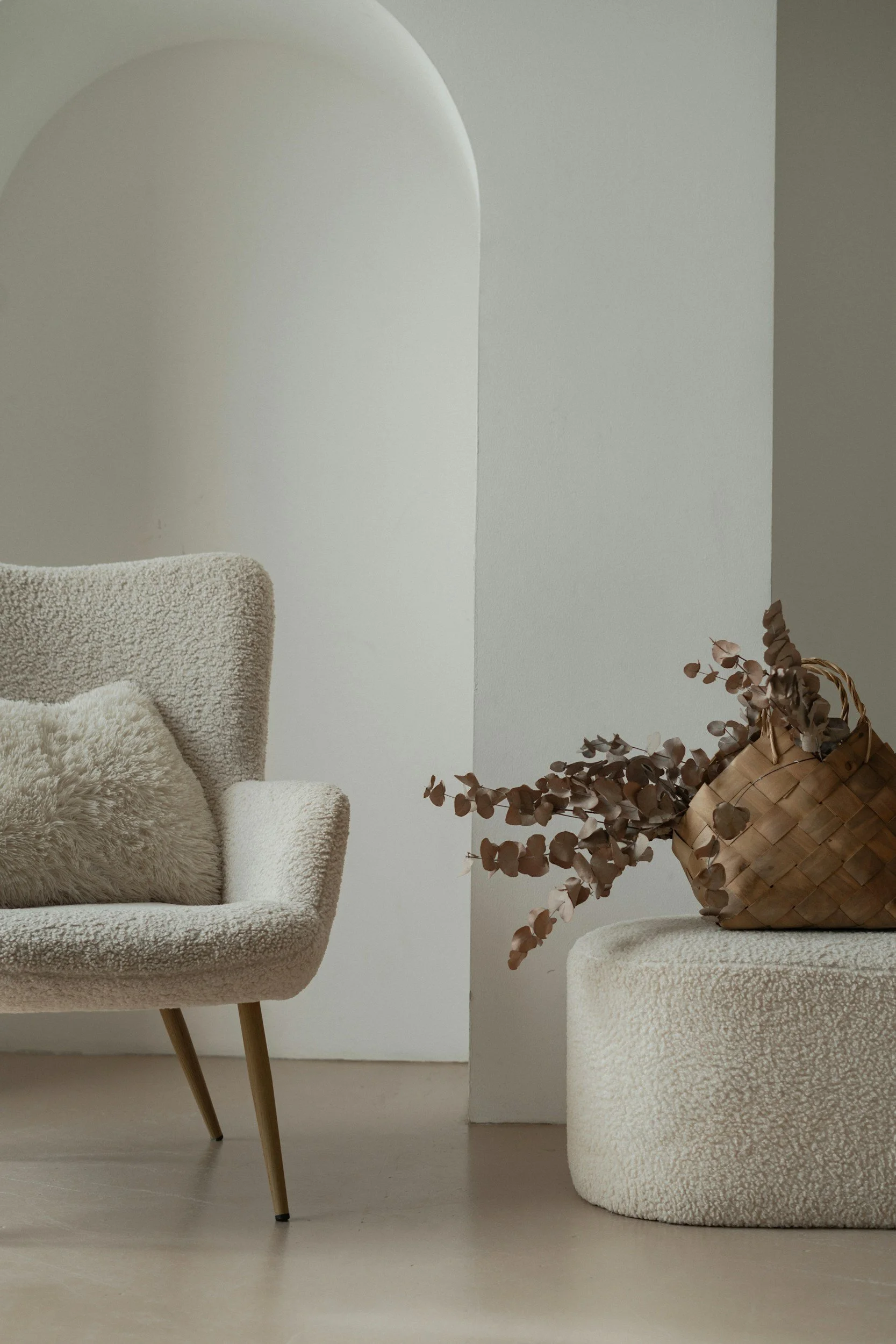 A beige upholstered armchair with wooden legs and a fluffy cushion, next to a white stool and a woven basket with dried eucalyptus branches, set against a plain light-colored wall.