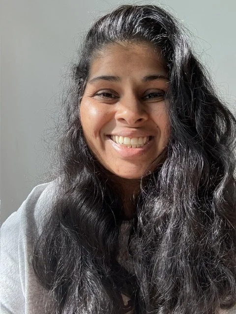 A young woman with long, dark, wavy hair smiling at the camera against a neutral background.