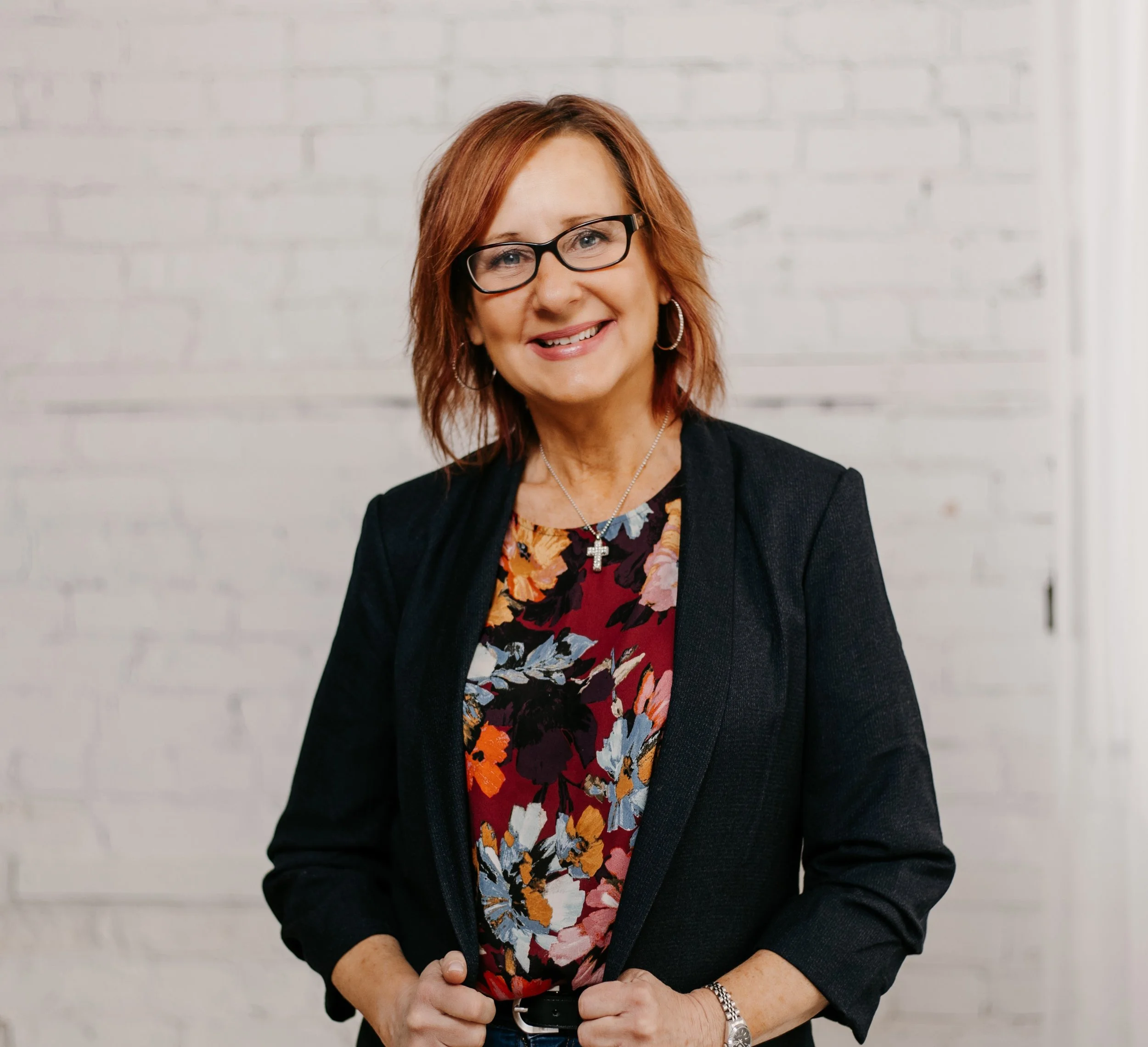 A woman with glasses and shoulder-length reddish-brown hair smiling at the camera, wearing a black blazer over a floral blouse, with a cross necklace and hoop earrings, standing in front of a white brick wall.