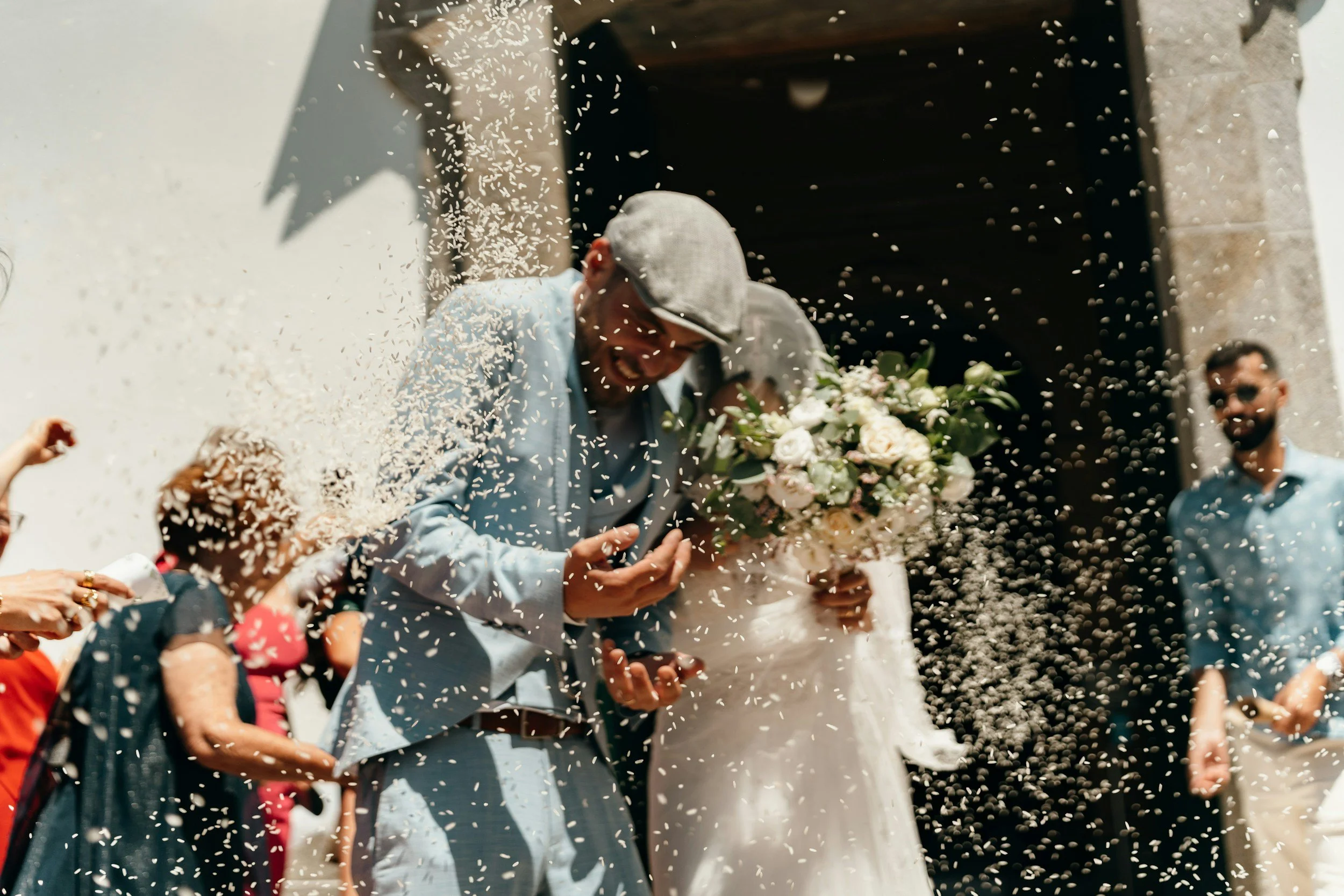 People celebrating a wedding outside, with rice being thrown at the couple. The groom is wearing a light blue suit and hat, while the bride is in a white wedding dress holding a bouquet. There are onlookers around them.