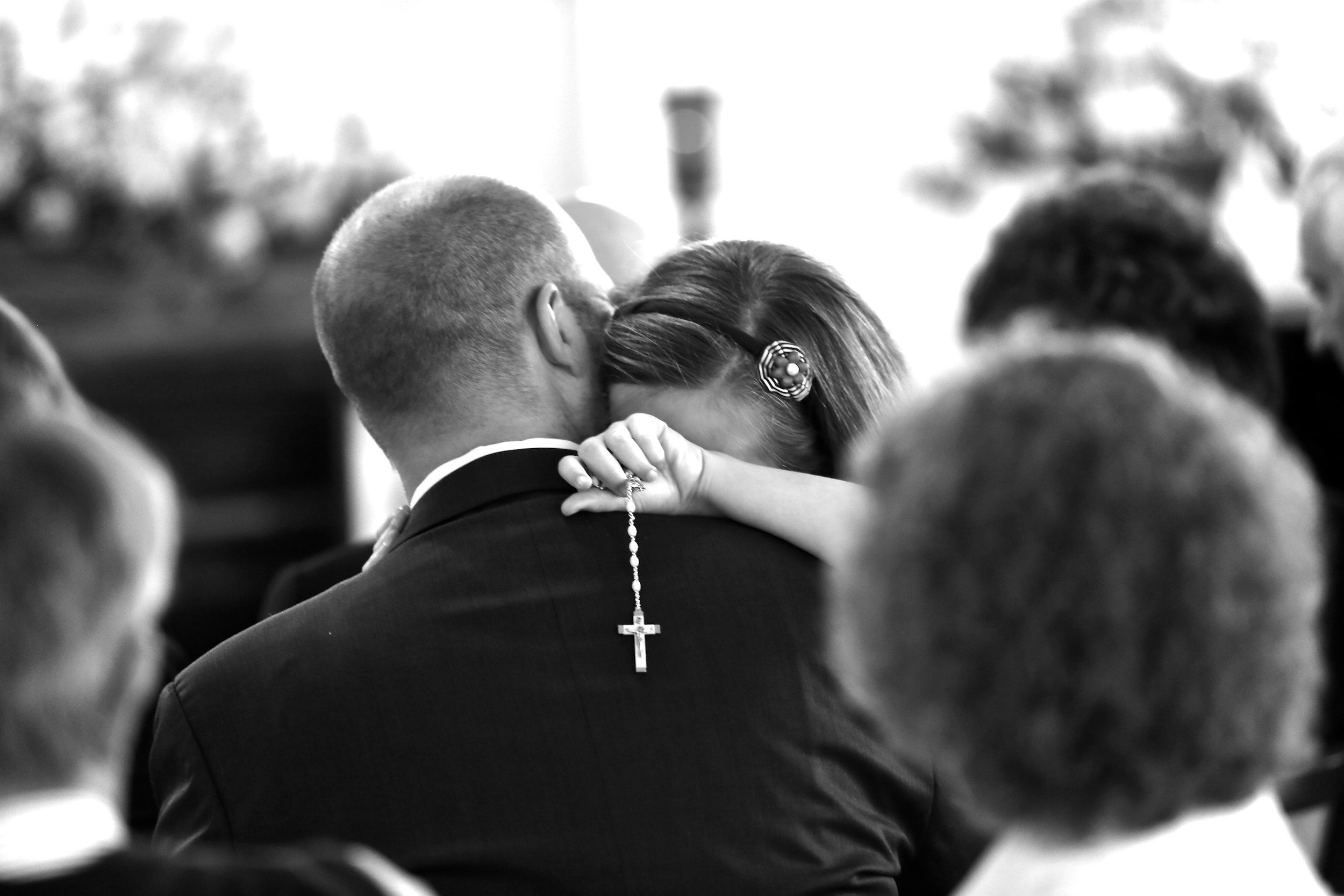 A woman leaning on a man's shoulder during a wedding ceremony. The man is wearing a suit with a rosary hanging from his collar. The woman is wearing a hairband with a decorative bow or flower. The image is in black and white, and there are other people visible in the background.