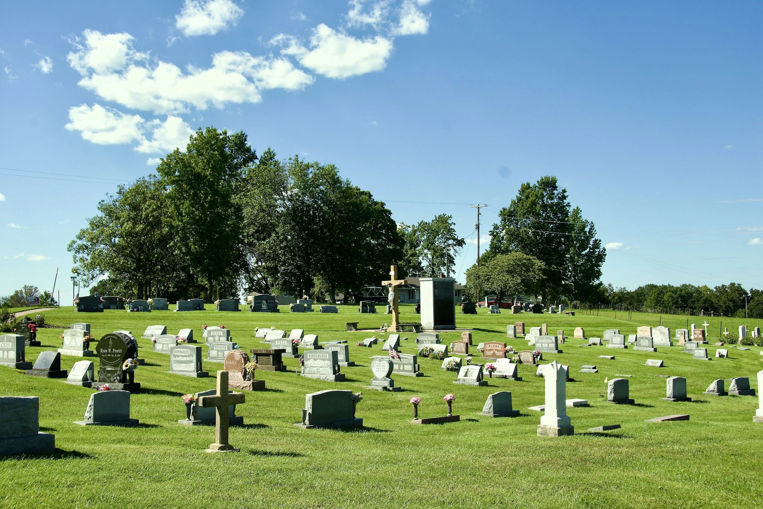 A cemetery with numerous gravestones, some with flowers, under a bright blue sky with scattered clouds, and a large cross in the background surrounded by trees.