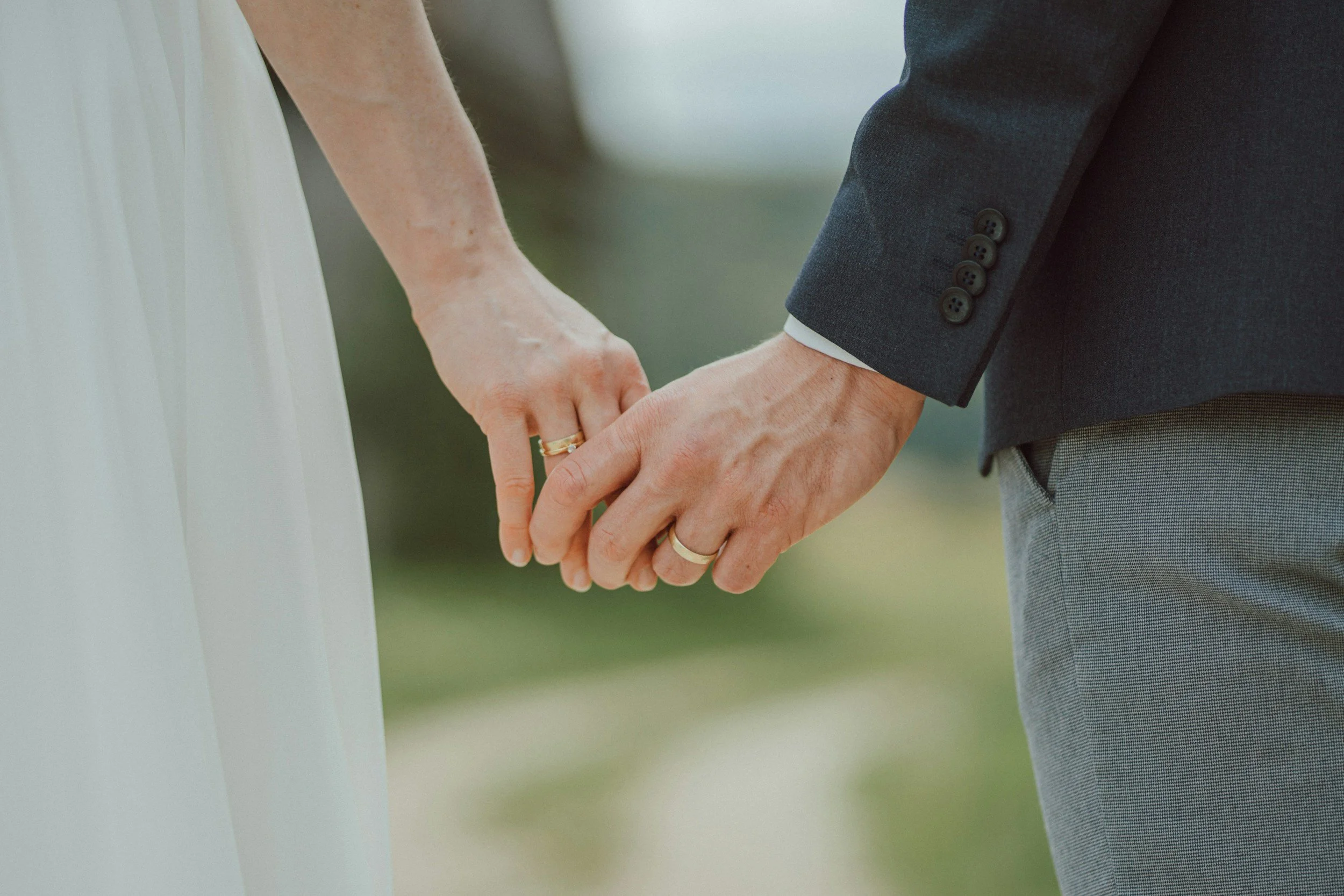 Close-up of a couple holding hands during a wedding, with the bride wearing a white dress and the groom in a dark suit, both with wedding rings.