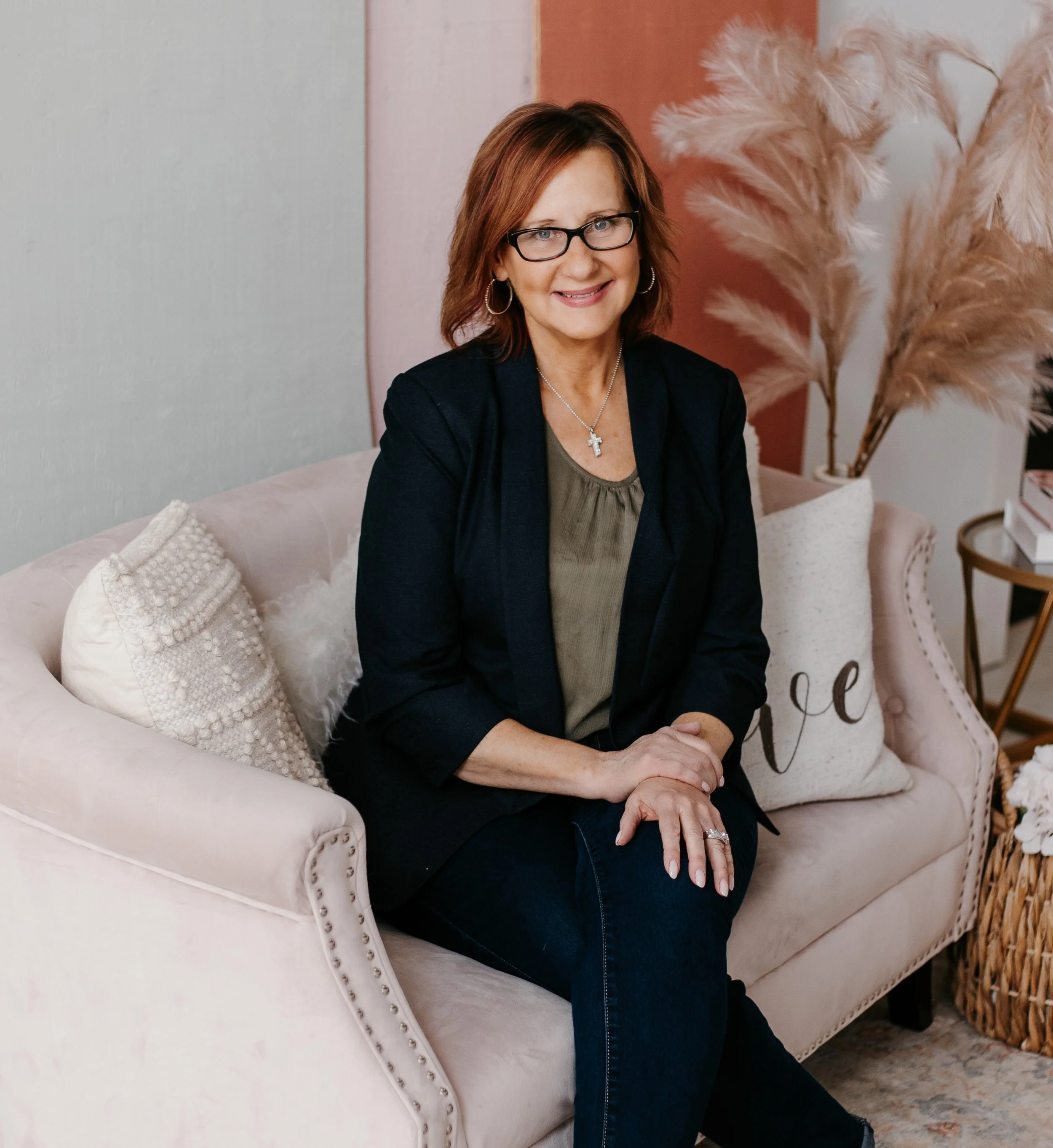 A woman with glasses and shoulder-length brown hair sitting on a light-colored sofa with decorative pillows, smiling at the camera, in a cozy living room with pampas grass in a white vase and a side table with books in the background.