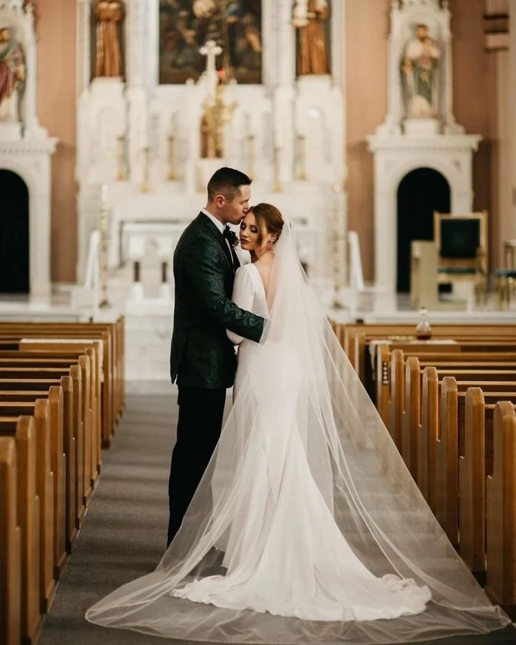 A bride and groom embracing in a church aisle during their wedding ceremony, with ornate altar and religious artwork in the background.