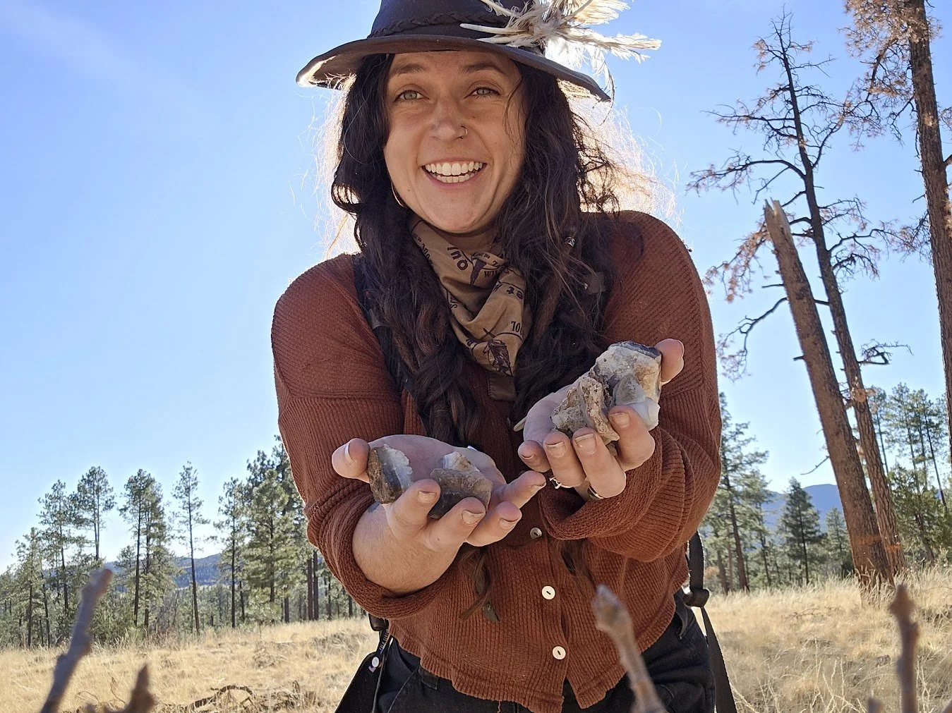 A woman outdoors in a forest holding rocks and smiling at the camera, wearing a hat with feathers, a brown jacket, and a bandana around her neck.