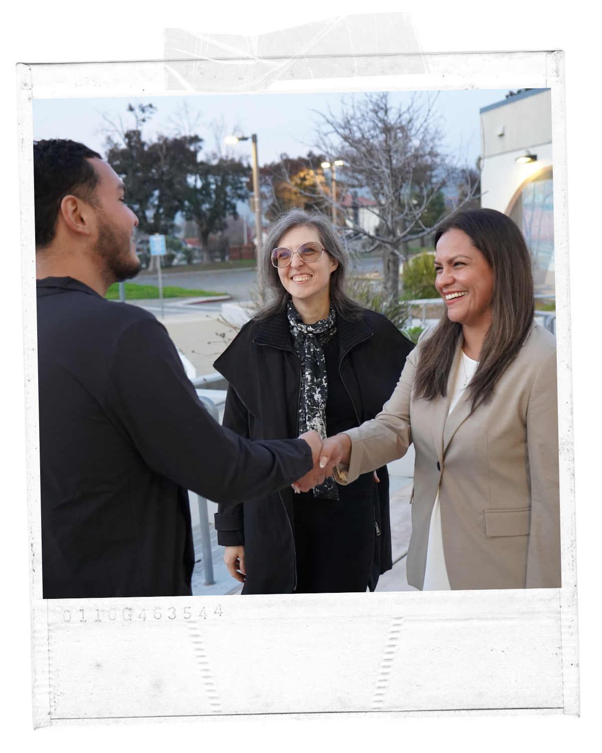 Three people outdoors, two women and a man, shaking hands and smiling, as a third woman witnesses. It is late afternoon, with trees and buildings in the background.