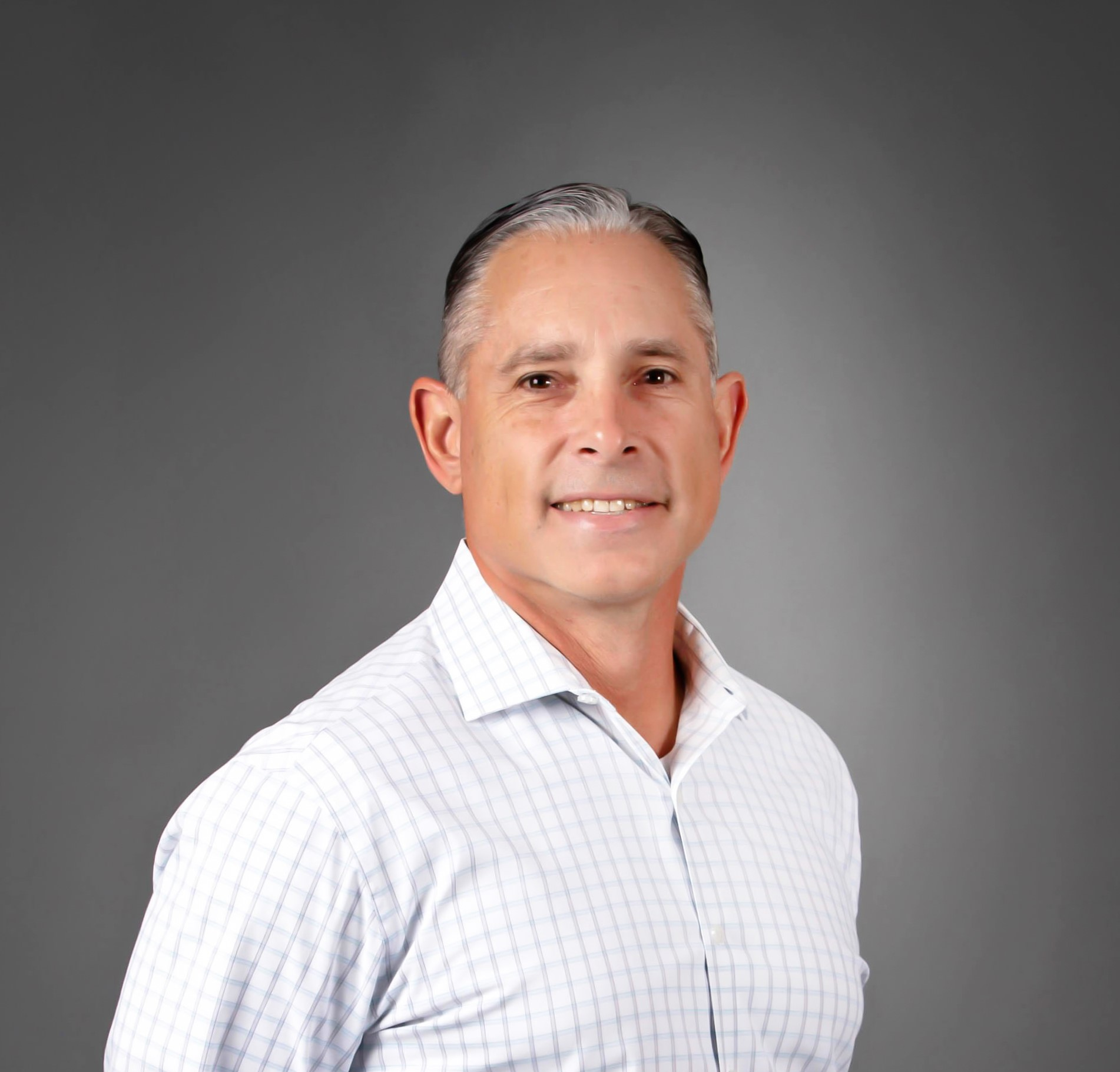 Portrait of a middle-aged man with short, slicked-back gray hair, wearing a white checkered dress shirt, smiling against a gray gradient background.