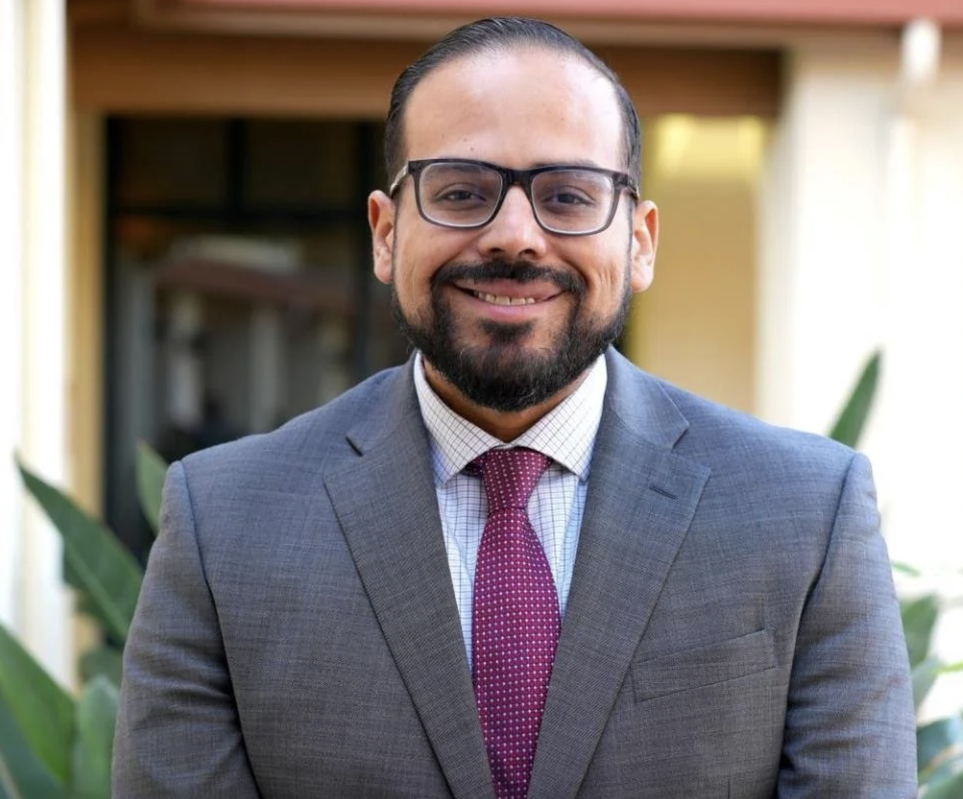 A man with glasses and a beard is smiling, wearing a gray suit, white checkered shirt, and a maroon tie, standing outdoors with green plants and a building in the background.