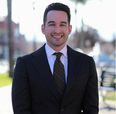 Smiling man in a black suit and tie standing outdoors on a sunny day.