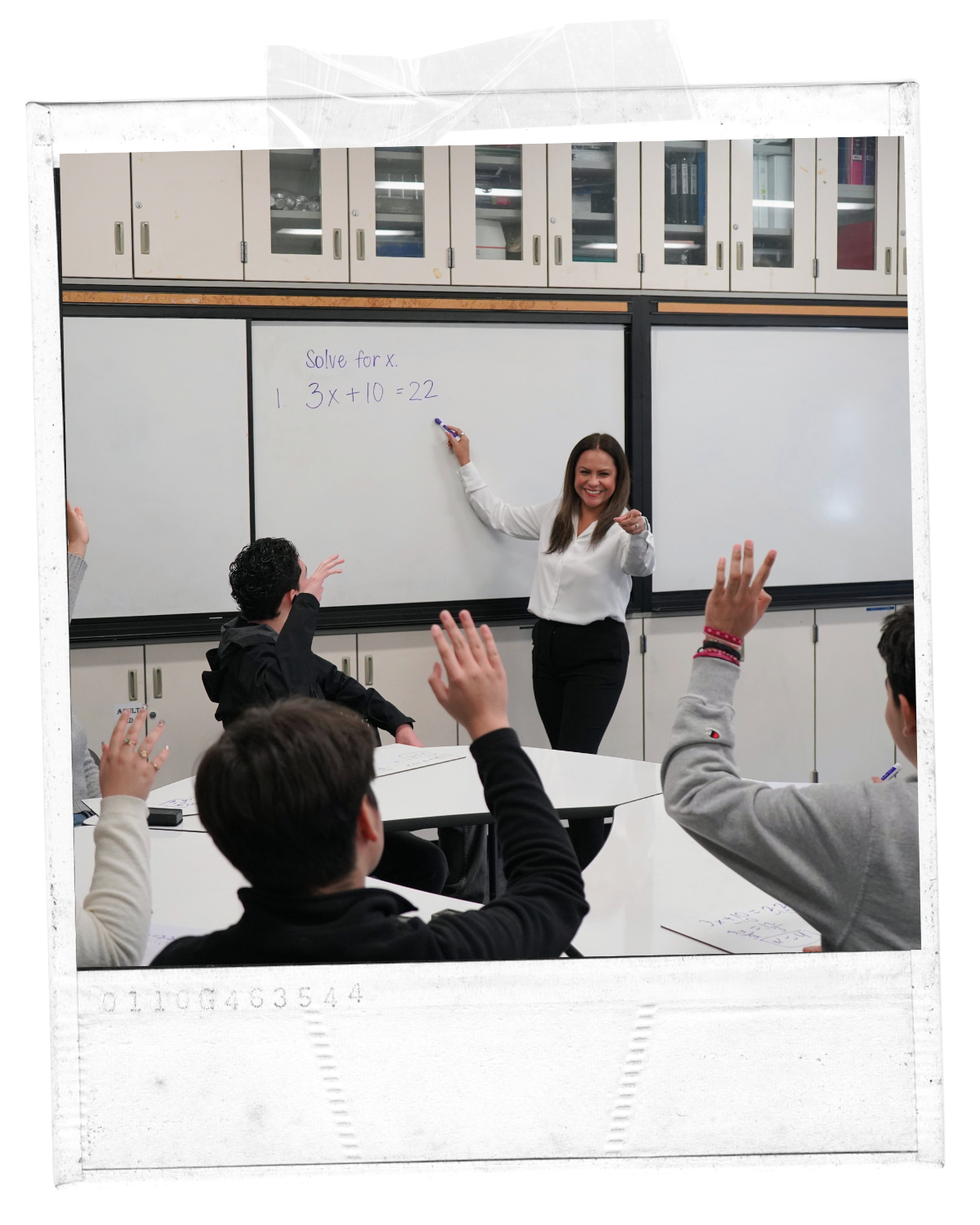 A classroom scene with a smiling woman teacher standing in front of a whiteboard with a math problem written on it. Several students are seated, raising their hands.