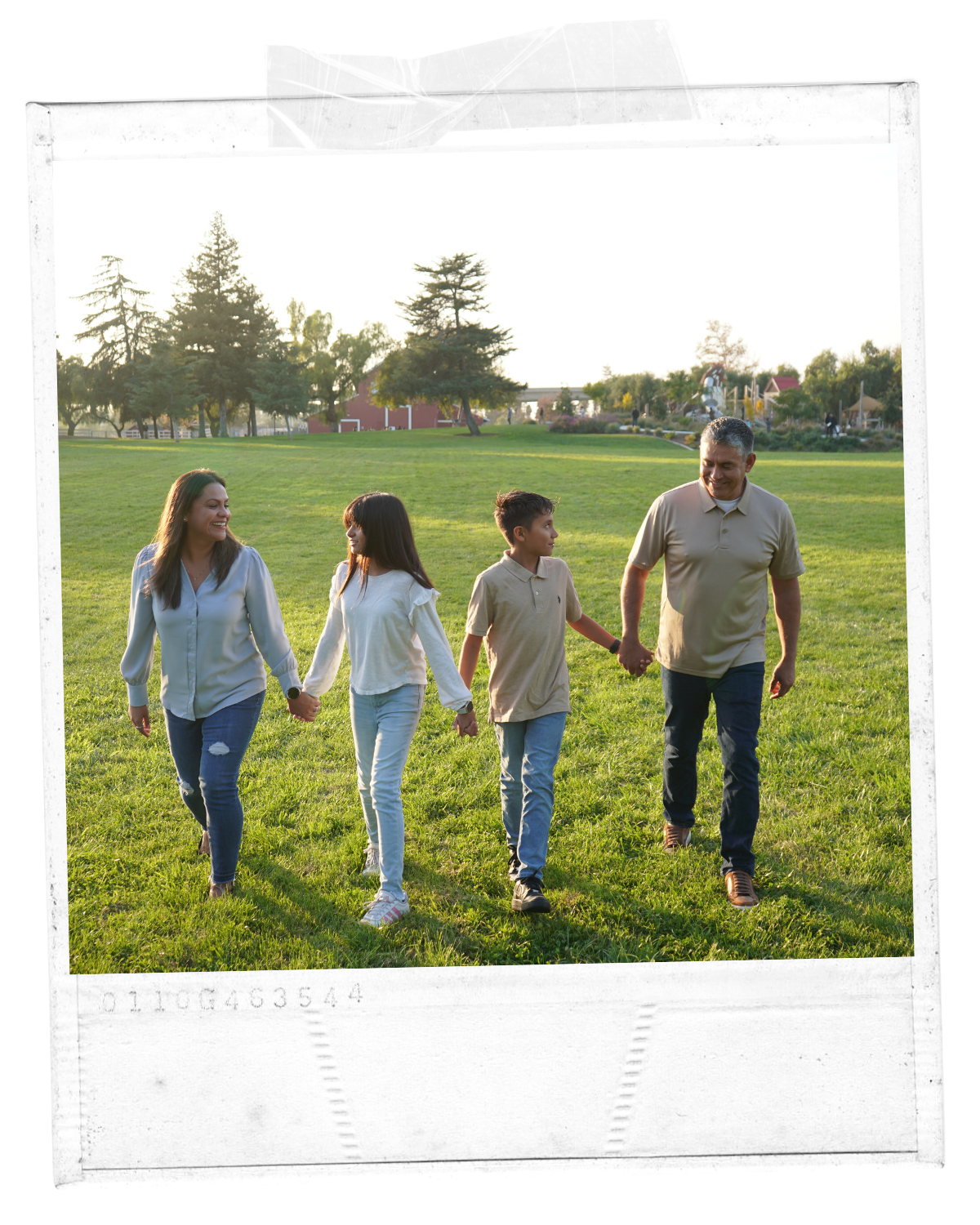 A family of four holding hands and walking across a grassy park area during sunset