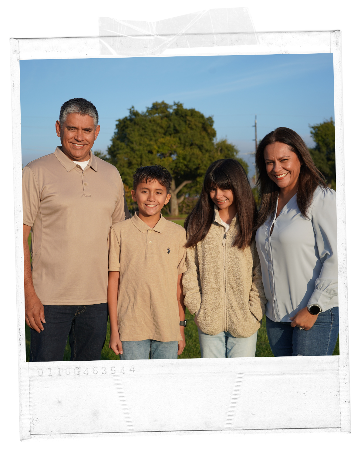 Family of five, two adults and three children, standing outdoors on grass, smiling, with trees and blue sky in the background.