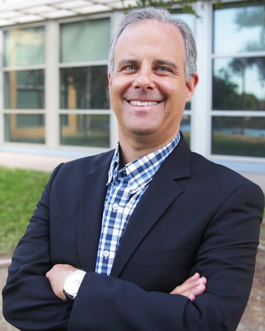 A smiling middle-aged man with gray hair, wearing a black blazer over a blue checkered shirt, standing outdoors in front of a modern building with large windows.