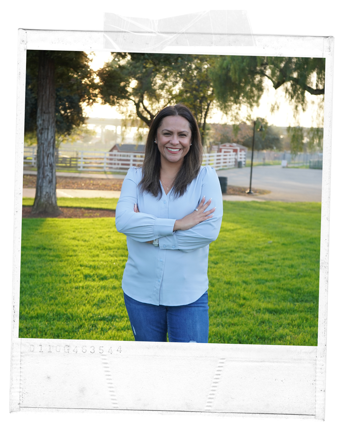 A woman standing outside on a grassy lawn during sunset, smiling with arms crossed, wearing a light blue blouse and jeans, with trees and a fence in the background.