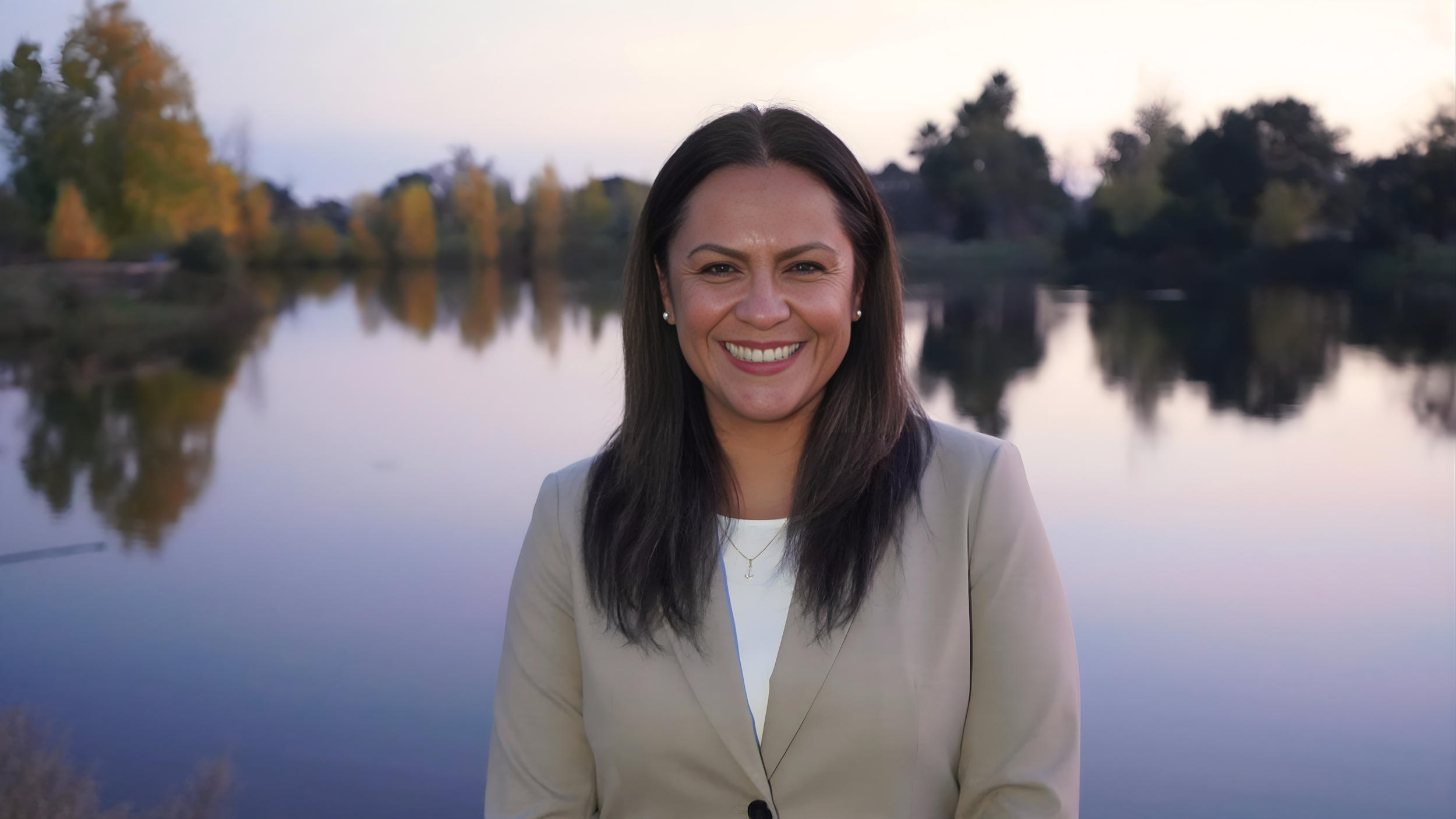 A woman smiling outdoors near a calm lake with trees and a soft sky in the background.