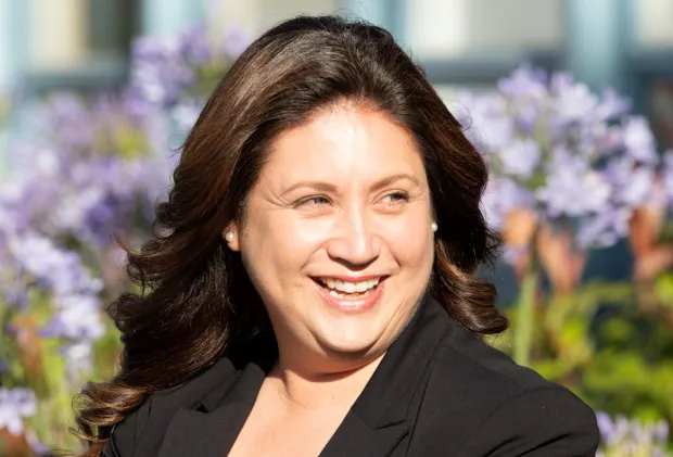 A woman with dark brown hair smiling outdoors with purple flowers in the background.