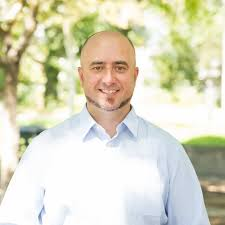 Portrait of a smiling man in a light blue shirt outdoors with trees in the background.