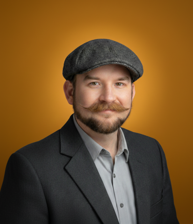 A man with a beard, styled mustache, and light eyes, wearing a gray newsboy cap and a dark suit with a light gray shirt, posing against a warm, orange-brown background.