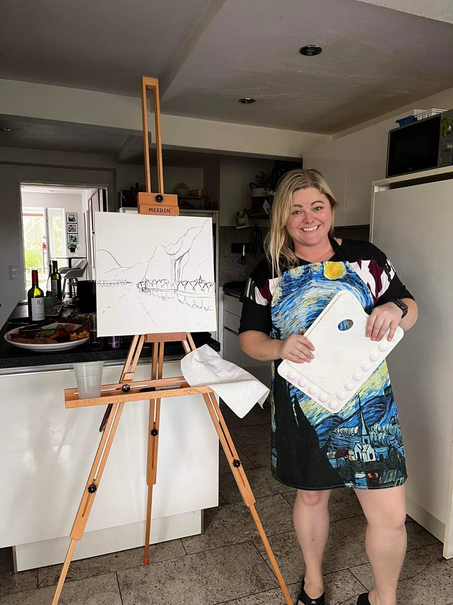A woman with blonde hair in a black dress and colorful apron holding a palette and paintbrushes, standing next to a blank canvas on an easel in a kitchen.