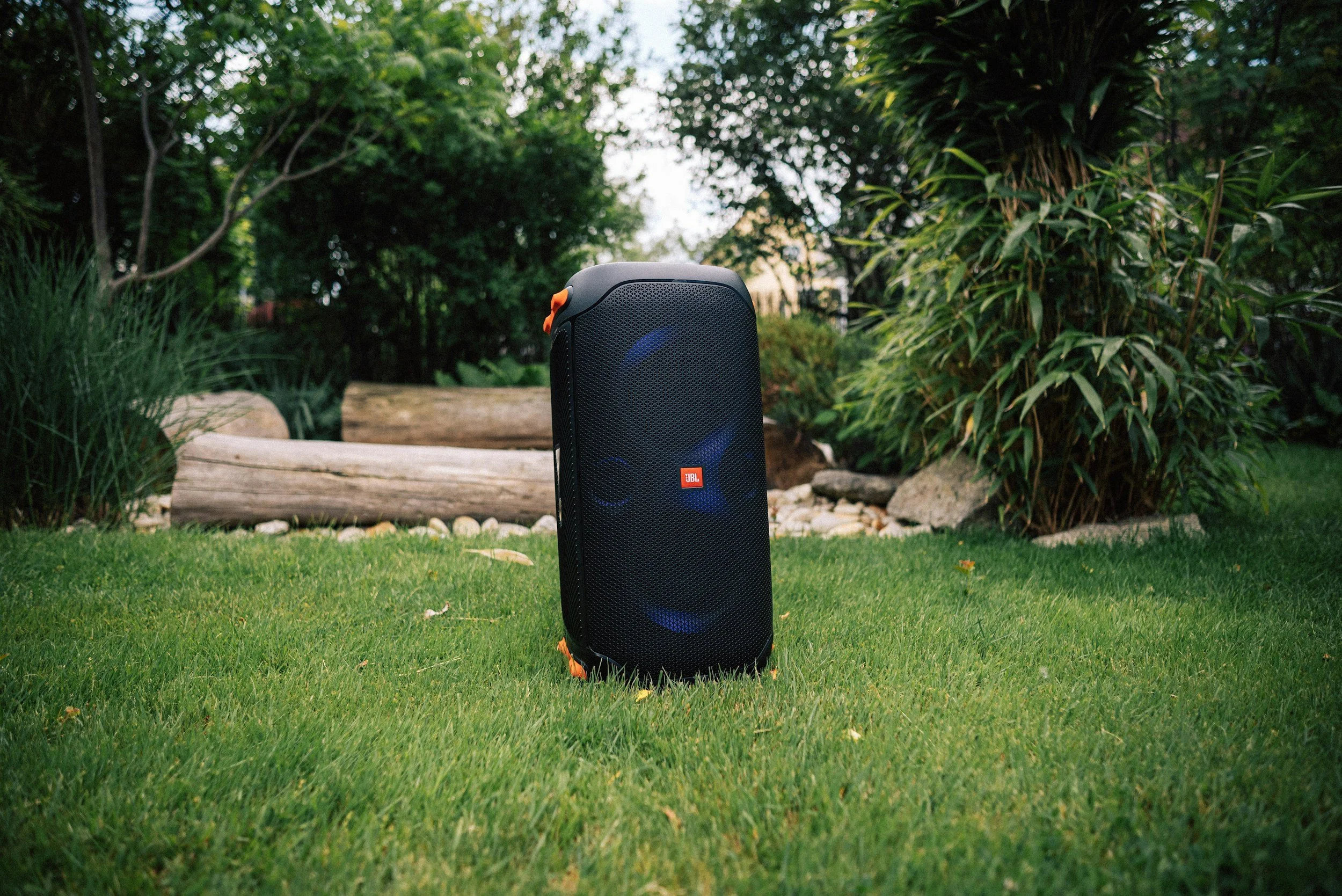 Outdoor scene with a black JBL portable speaker on a grassy lawn, surrounded by trees, bushes, and rocks.