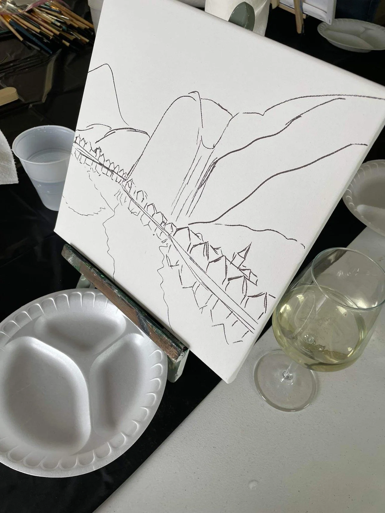 A painting in progress on a canvas shows a landscape with mountains, trees, and a road. Art supplies and a glass of white wine are on the table.