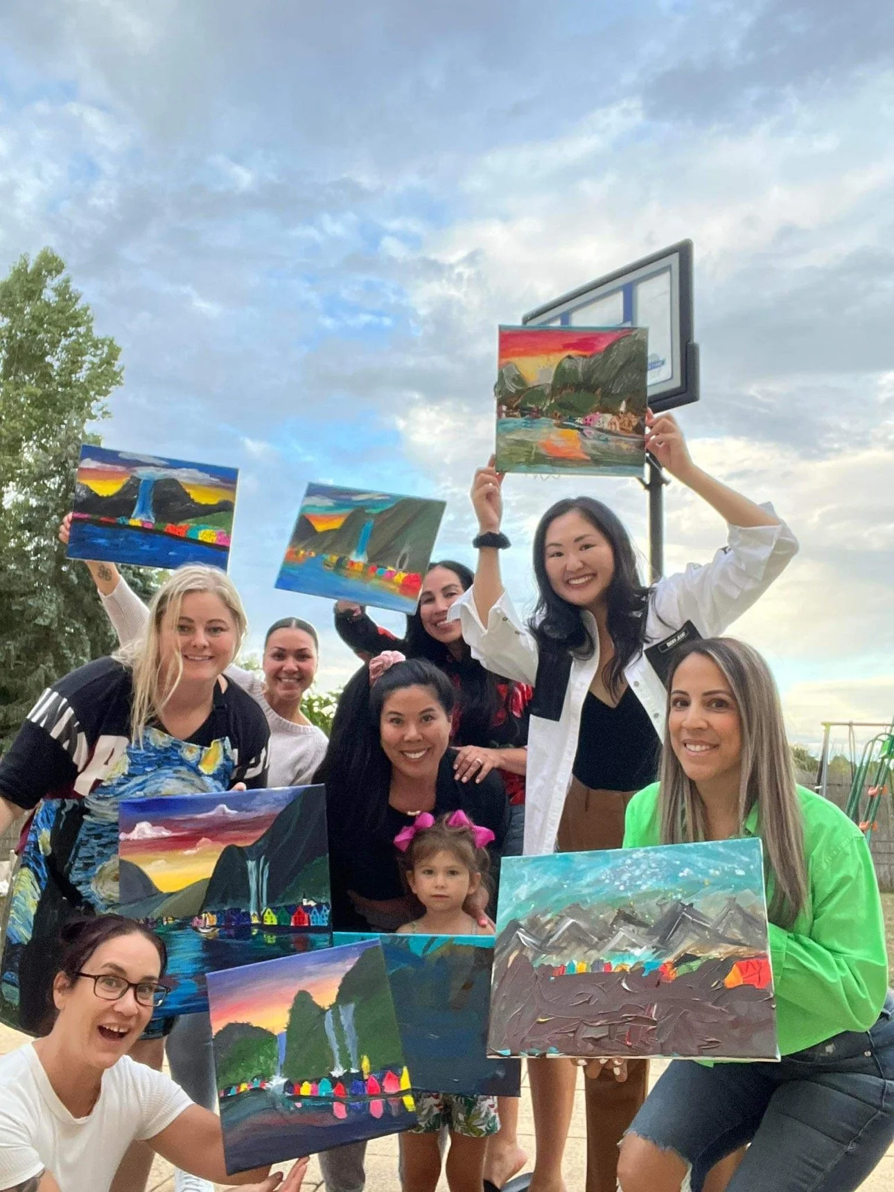Group of women and a young girl outdoors, holding colorful landscape paintings, smiling under a partly cloudy sky.