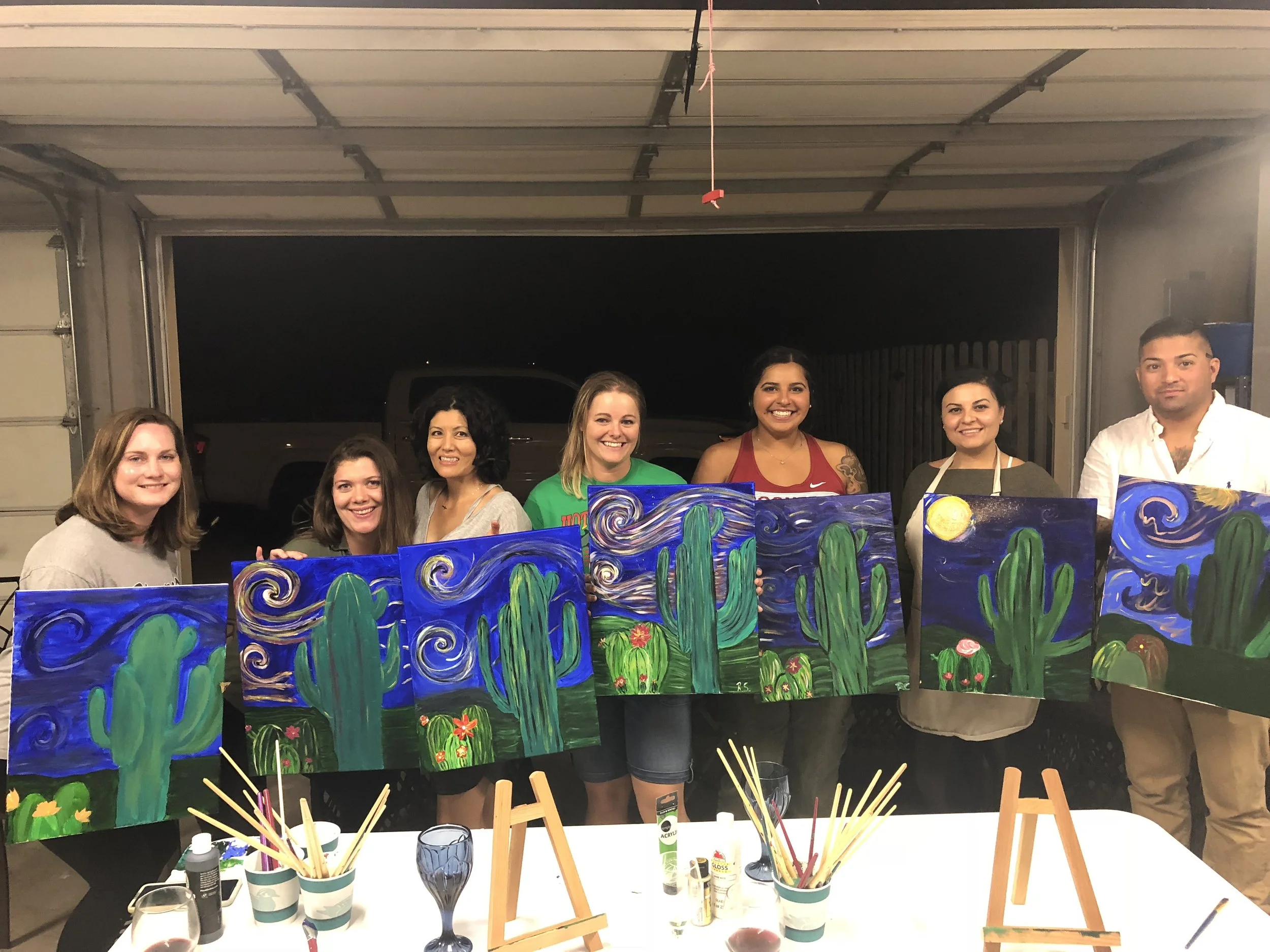 Group of seven people standing in a garage, holding up paintings inspired by Van Gogh's Starry Night featuring cacti and desert landscape.