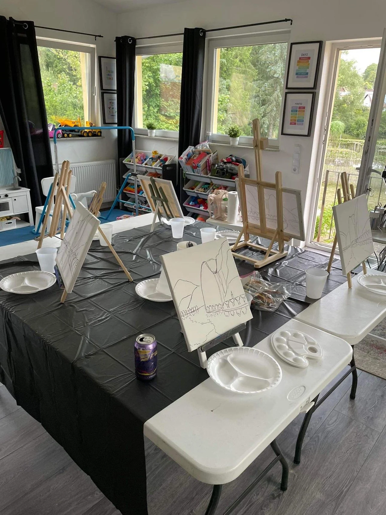 Indoor art studio with a large black table covered in art supplies, easels with partially completed paintings, paper plates, and a soda can. The room has large windows with black curtains and shelves filled with art materials.
