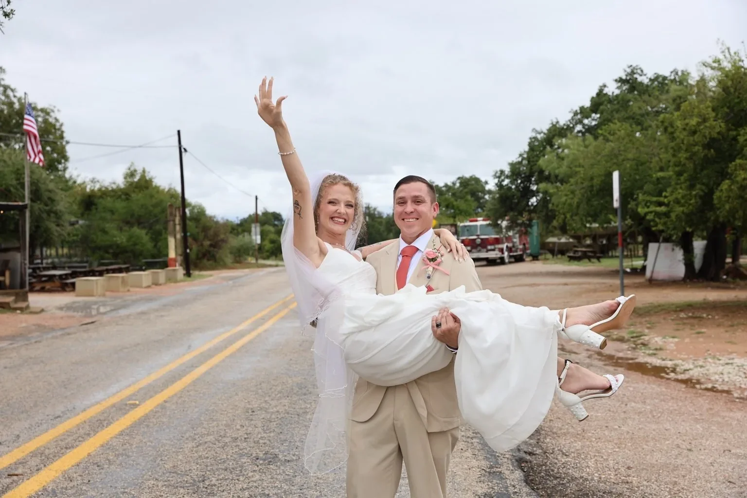A smiling groom carries the bride as she raises her hand in celebration after their wedding.