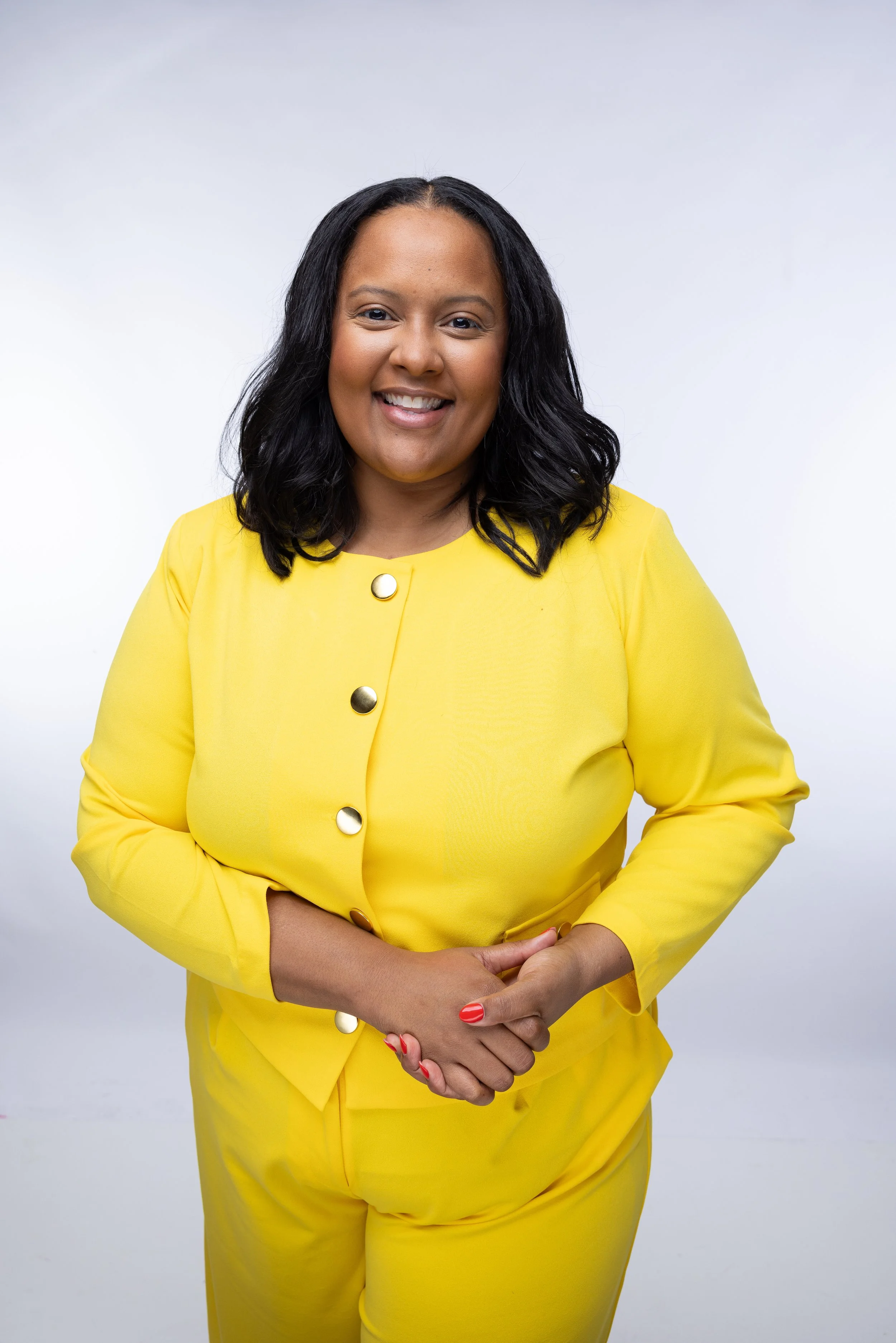 A smiling woman with black hair in a yellow suit with large gold buttons, standing against a white background.