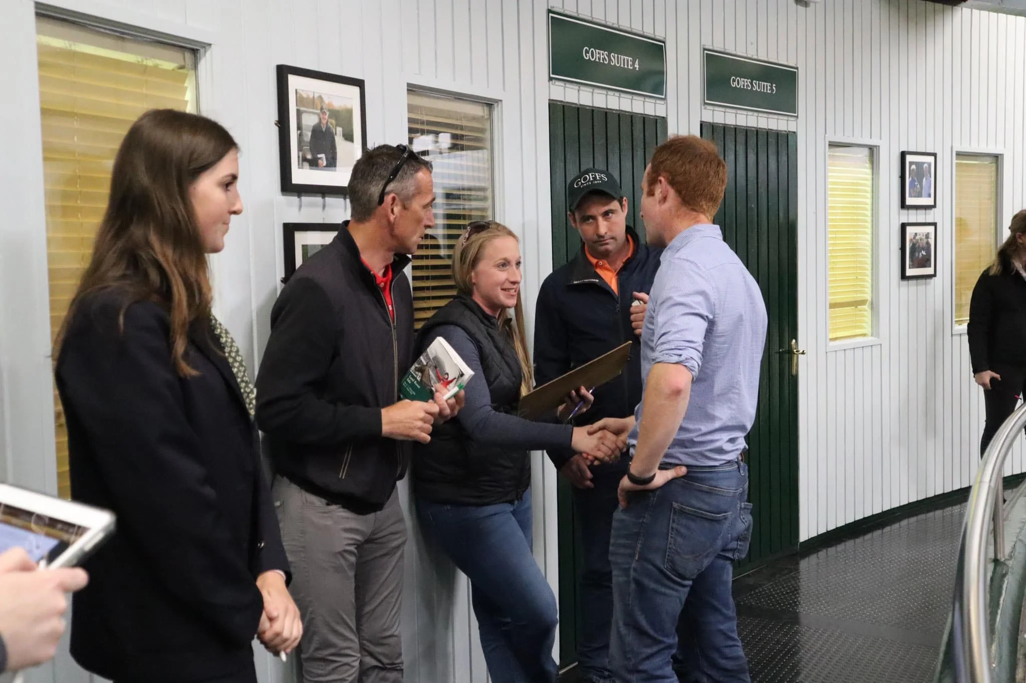 Group of people standing in line, shaking hands and conversing in an indoor setting with framed photographs on the wall and signs labeled 'GOFFS SUITE 4' and 'GOFFS SUITE 5'.