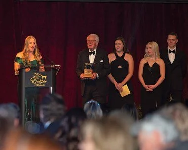 A woman speaking at a podium during an award ceremony, with four people standing on stage behind her, including a man holding an award, all dressed formally.