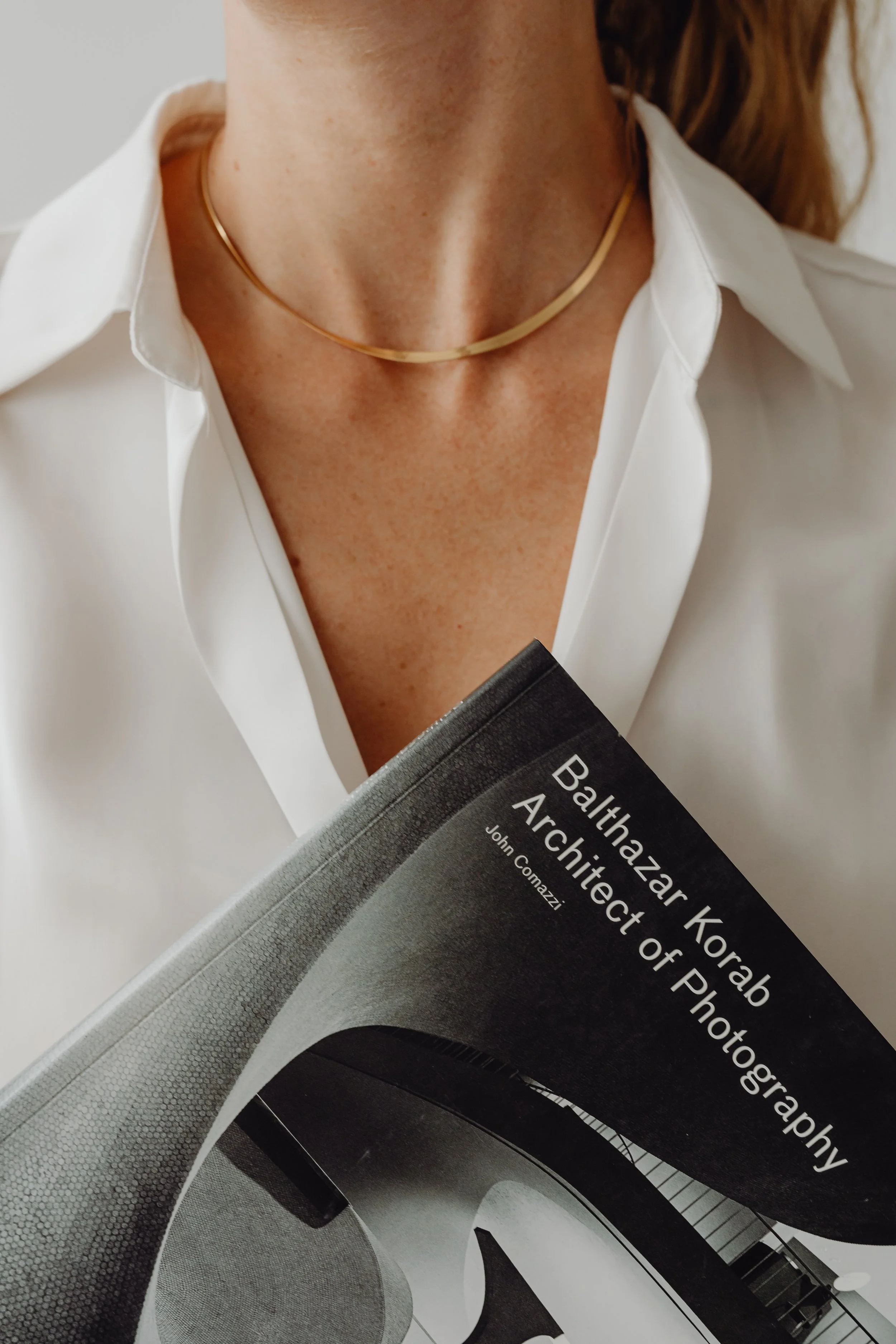 A woman wearing a white blouse and a gold necklace is holding a book titled "Balthazar Korab: Architect of Photography" by John Comazzi, with her face not visible.