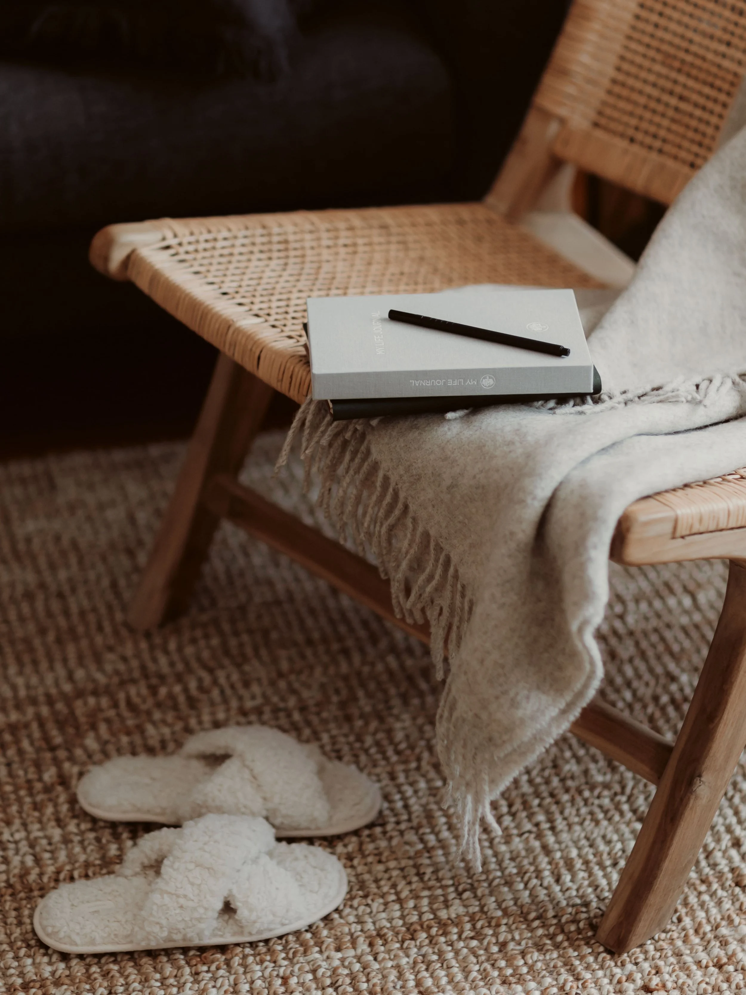 A cozy scene with a woven chair, a soft blanket, a notebook, and slippers on a textured rug.