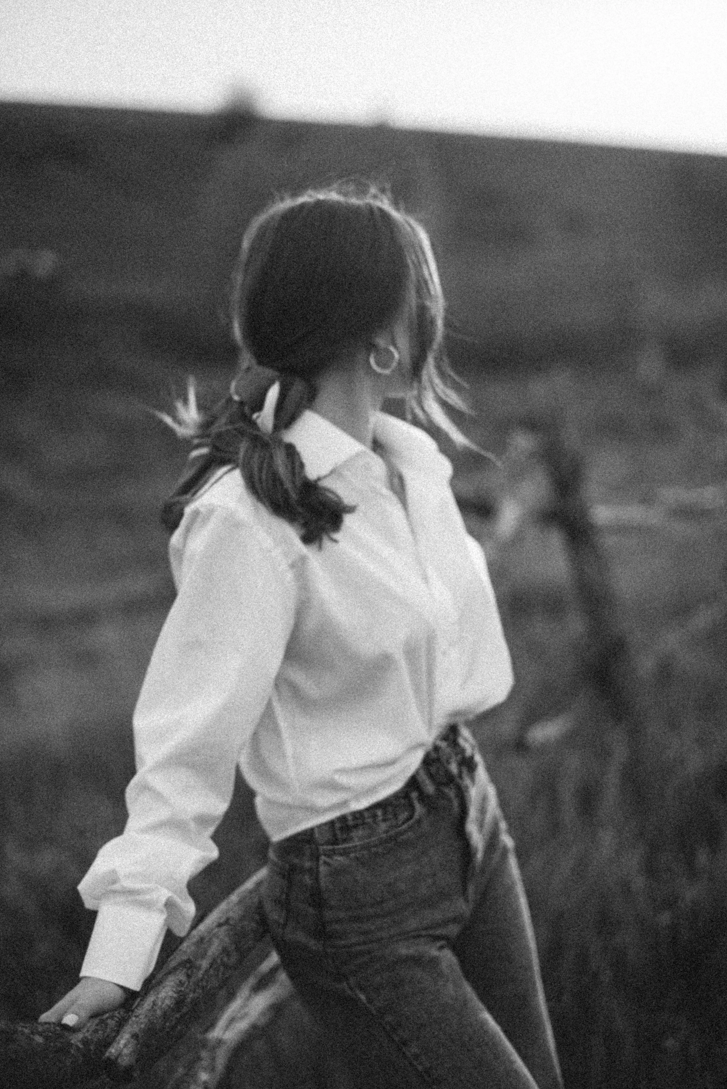 A woman with tied-back hair, wearing a white blouse and jeans, leaning against a fallen tree trunk outdoors in a natural setting, with a blurred figure in the background.