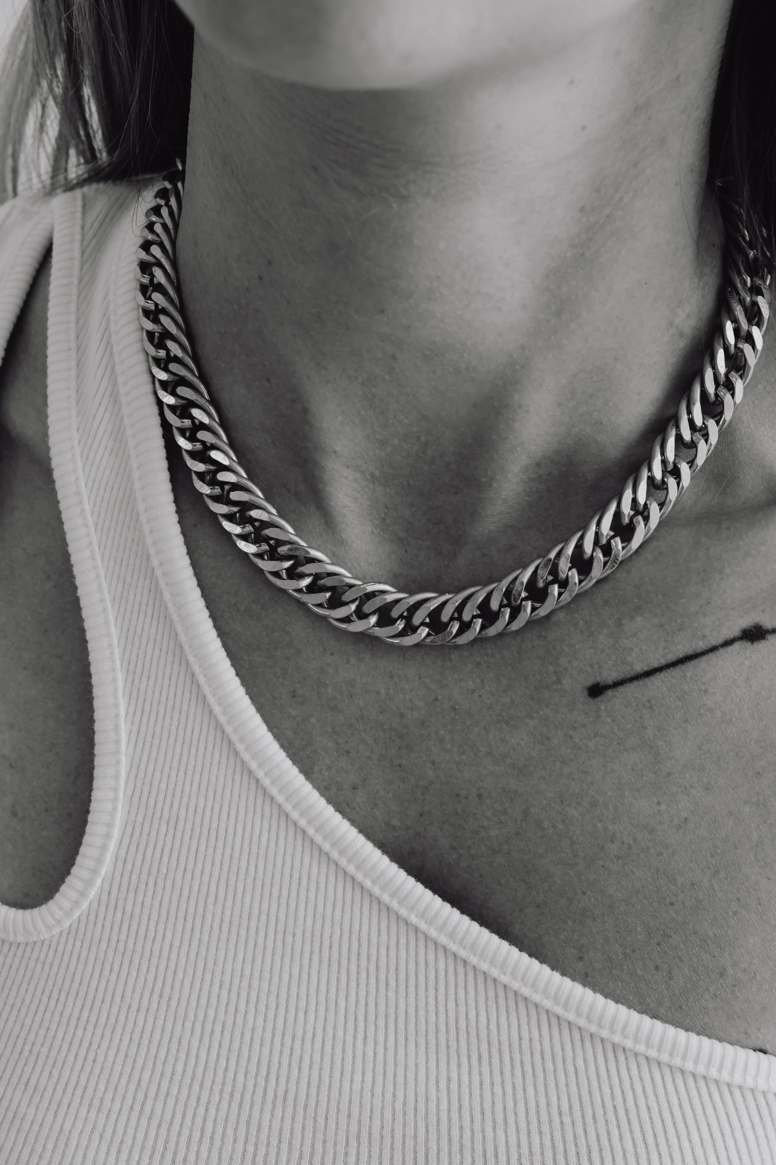 Close-up of a person's neck wearing a thick silver chain necklace, with a tattoo on the upper chest, dressed in a white tank top.