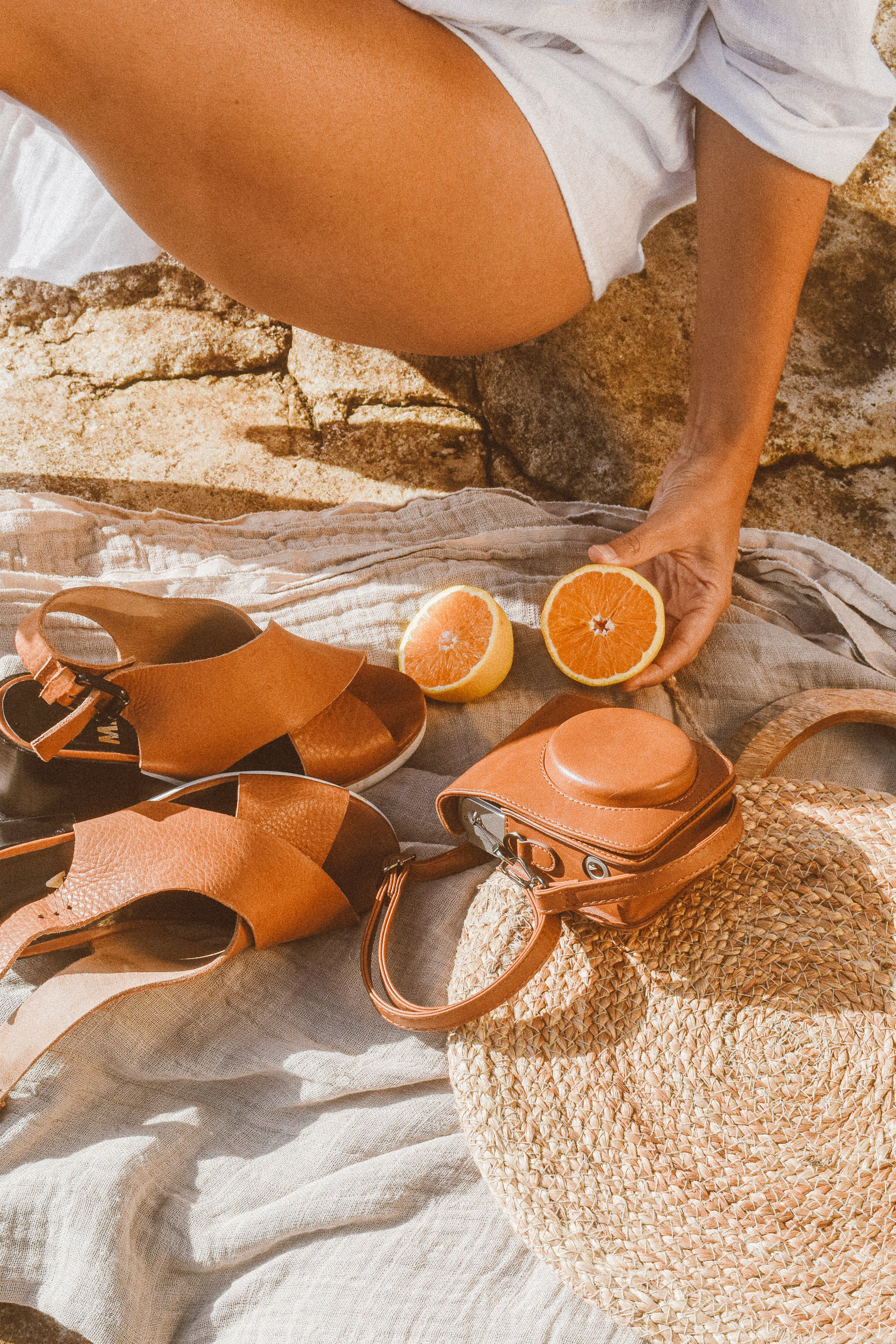 A person's hand holds a halved orange on a light-colored fabric with sandals, a straw bag, and an old camera arranged nearby, suggesting a beach or outdoor setting.