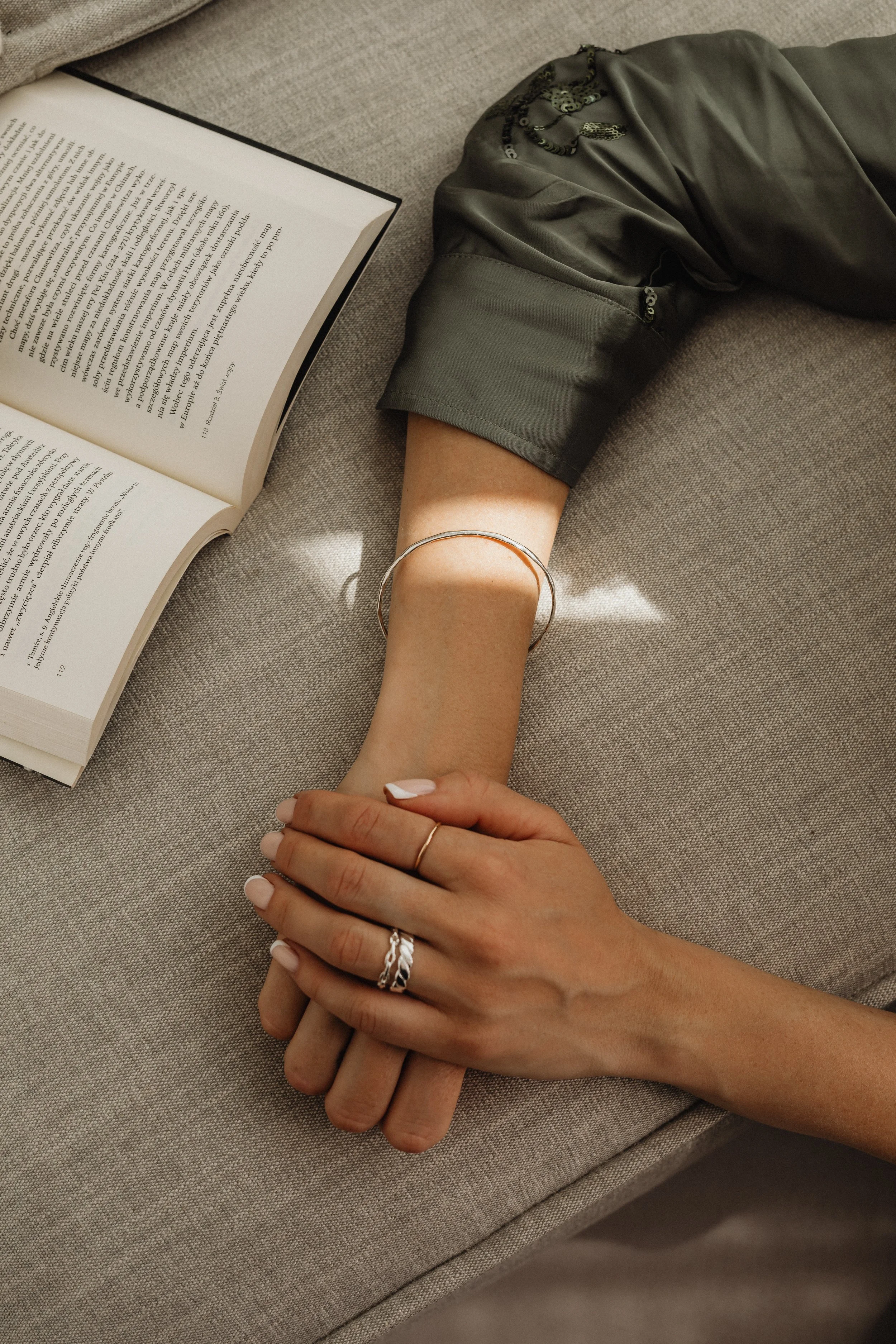 A person resting their hand on a beige fabric surface, wearing multiple silver rings and a silver bangle, with an open book nearby and sunlight casting a shadow.